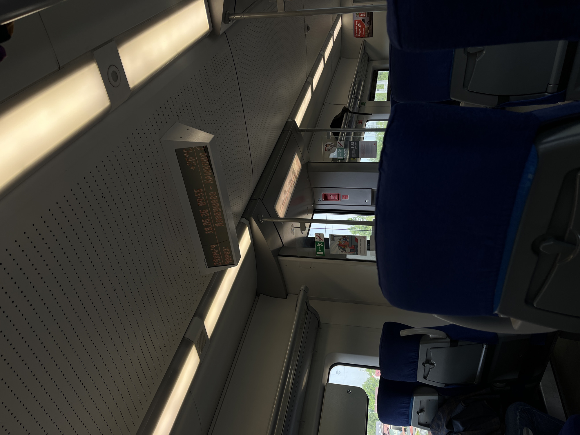 Here is a description of the image:
The image shows the interior of a train car. The ceiling is white with evenly spaced light fixtures. A display shows the date, time, temperature (26°C), and the station names "Alabushevo - Kryukovo". The speed is shown as 24 km/h. The seats are upholstered in blue. There is a window to the outside showing foliage. A sign with "100 рублей" is also visible. Based on the stations named on the display, this train is likely in or near Povarovo, Russia, as Alabushevo and Kryukovo are neighborhoods in the area of Zelenograd and located in the Moscow region.