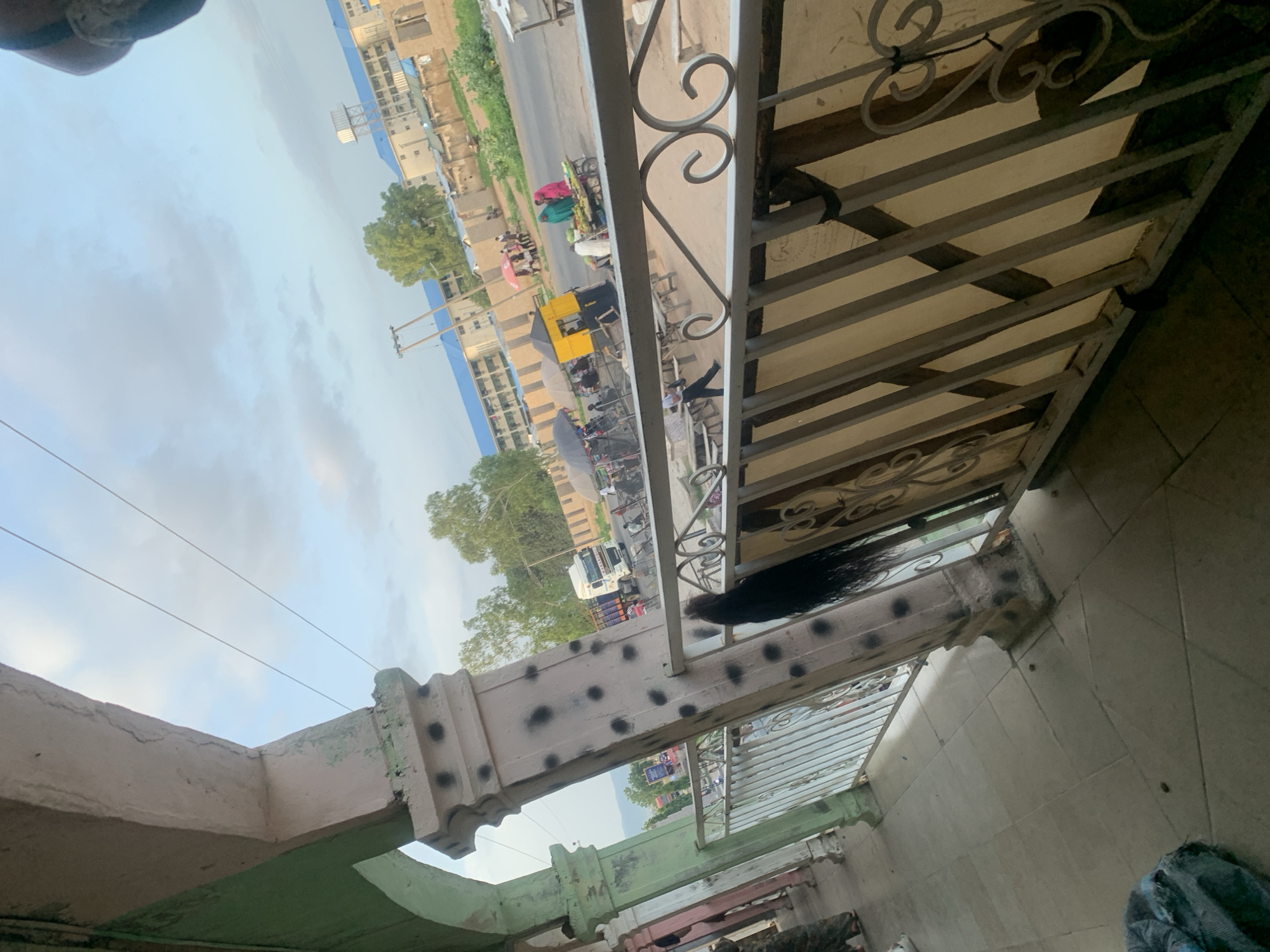 The image shows a street scene in Jalingo, Nigeria, viewed from an elevated vantage point, possibly a balcony or verandah. In the foreground, there is a tiled floor and a metal railing with decorative scrollwork. A large, dark, braided hairpiece is attached to one of the supporting pillars of the railing.
Beyond the railing, a street is visible with various activities. A white truck with "MAZAFAS" written on its side is driving on the road. There are several market stalls with umbrellas providing shade, and people are seen interacting. One stall has a yellow kiosk, and another features vendors with a cart laden with fruits. Some individuals are dressed in traditional Nigerian attire. In the background, buildings with blue roofs are visible, along with trees and a water tower. The sky is partly cloudy. The overall impression is of a bustling urban environment with a mix of commercial activity and everyday life.