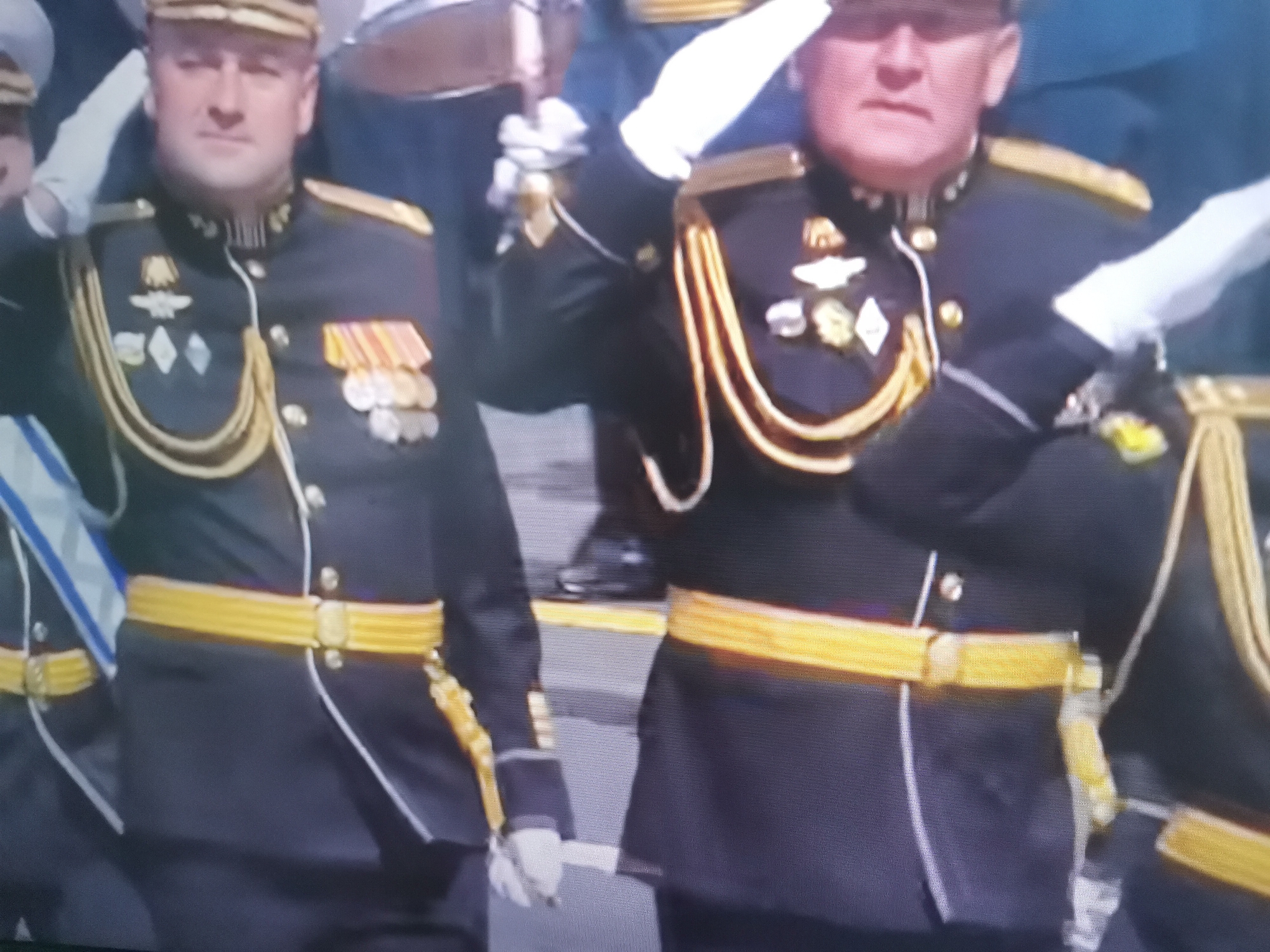 The image shows two men in ceremonial military uniforms, standing at attention. They are wearing dark blue jackets with gold trim, epaulets, and sashes. Both men have medals pinned to their chests and are wearing gold belts. The man on the left is wearing a peaked cap, and the man on the right appears to be wearing a similar cap. The background is blurred, but it appears to be an outdoor setting, possibly a parade or ceremony. The men's posture and attire suggest a formal occasion. The context of Belaazyorsk, Belarus, suggests this might be a military event or commemoration within that location. The image is captured from a slightly elevated angle, focusing on the upper bodies of the two individuals.