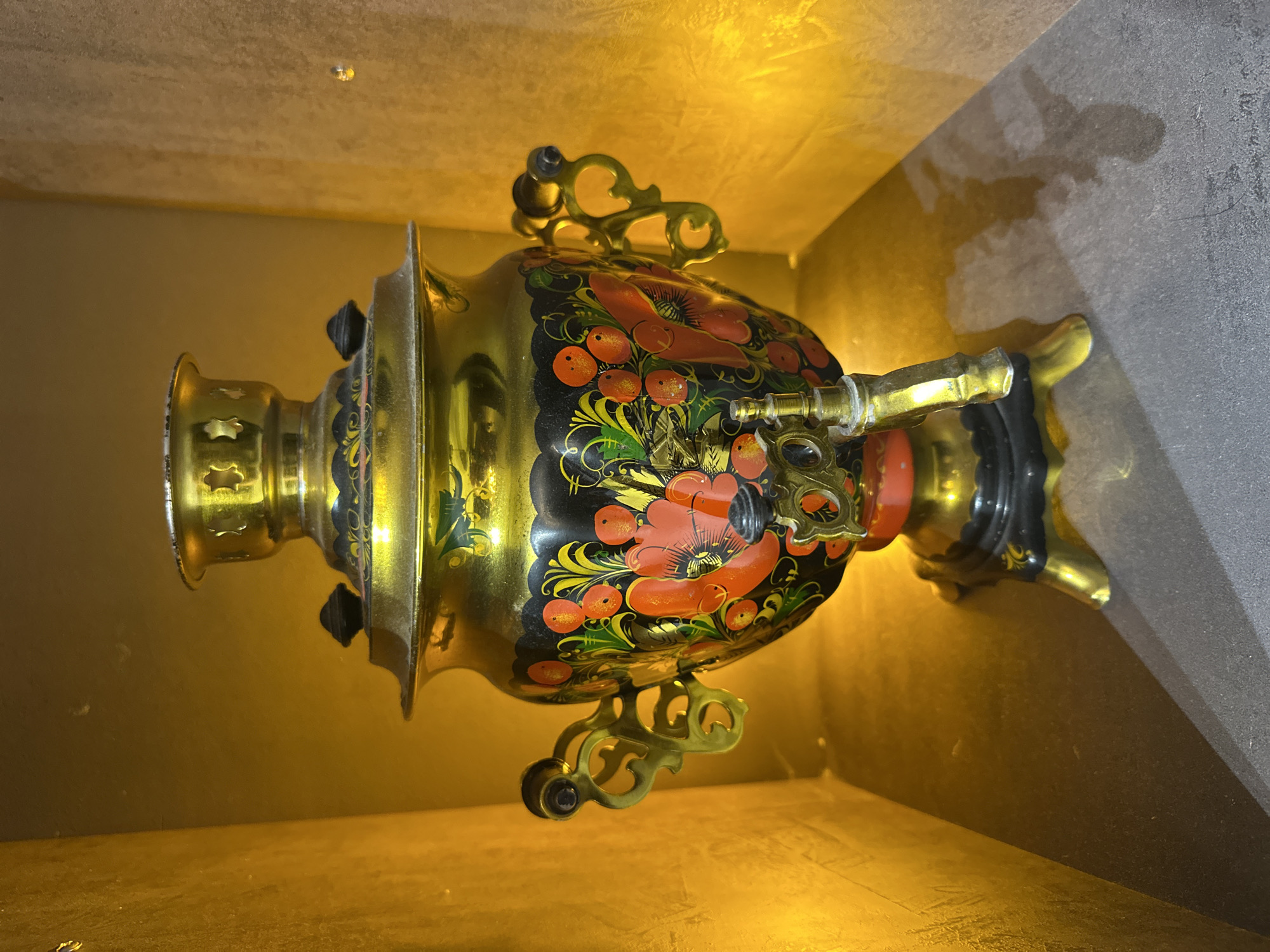The image shows a samovar, a traditional Russian metal container used to heat water, sitting in a niche. The samovar is primarily gold in color and decorated with a black and red floral pattern. It has ornate handles and a spout. The niche is illuminated with a warm, yellow light. The background is split, with one side appearing to be a gray wall and the other side is a gold colored wall. There are no people visible. The location, Kursk, Russia, is not explicitly indicated in the image but included based on the request.