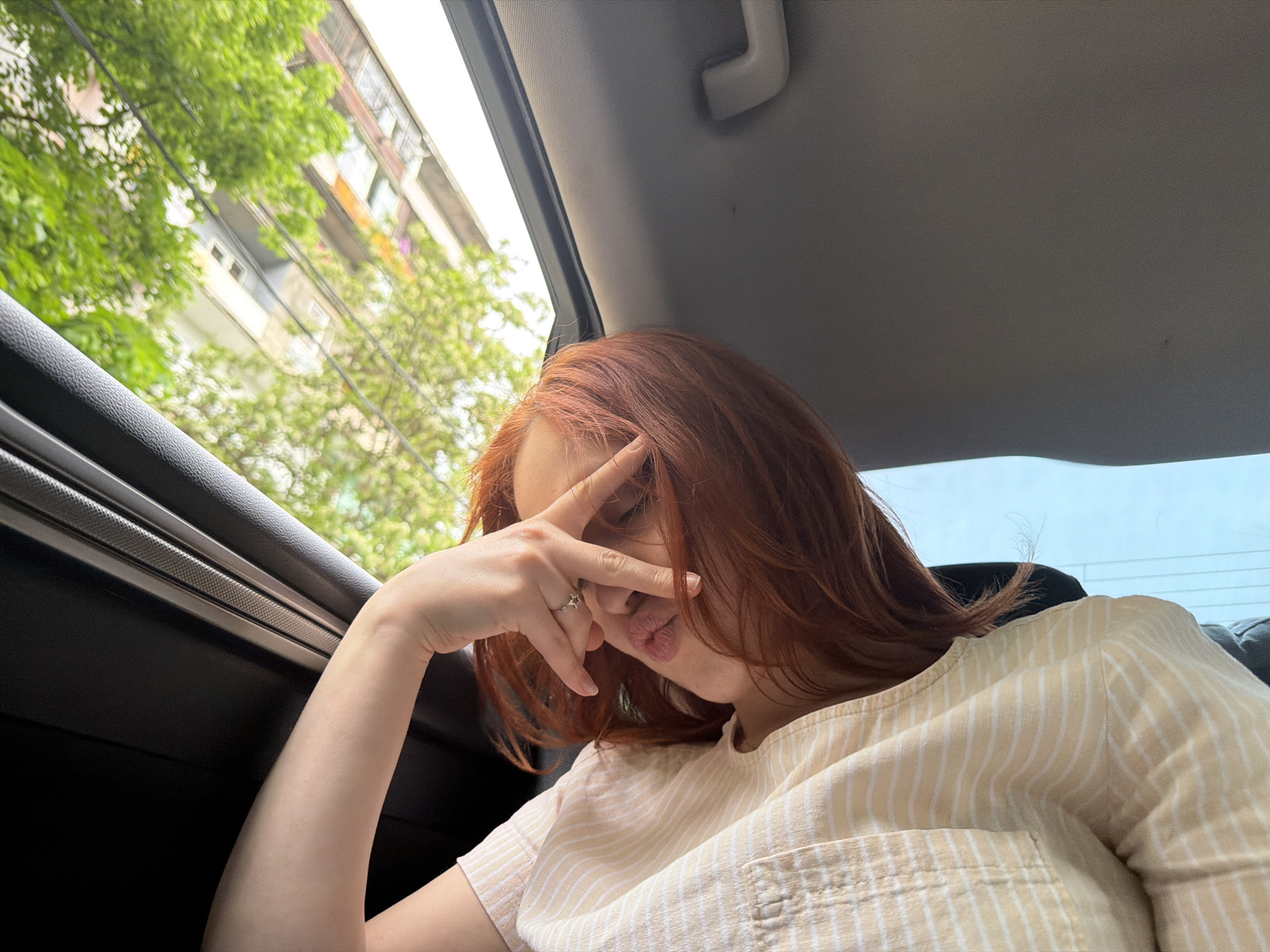 A woman with red hair is seated in the back seat of a car. She is wearing a light yellow and white striped shirt, and her hand is raised with her fingers spread across her forehead in a playful gesture. A silver ring is visible on her finger. The car window shows a cityscape with green trees and buildings, potentially located in Tbilisi, Georgia. The interior of the car includes a gray ceiling and dark window frame.