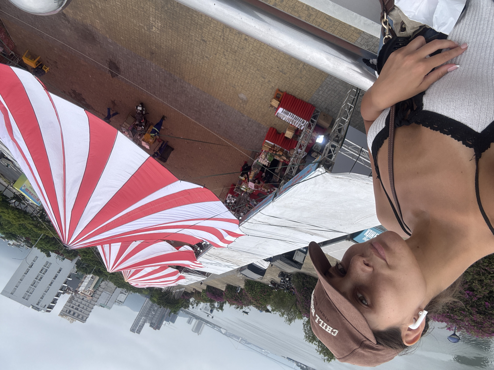 The image is taken from an elevated position in Đà Nẵng, Vietnam, overlooking a market area with red and white striped tents. The cityscape, including buildings and a bridge, is visible in the background. In the foreground, a person wearing a brown cap and earbuds is positioned to the right of the frame. The person is also wearing a white top with black trim. A silver railing is partially visible in the upper portion of the frame. The market area shows stalls and people.