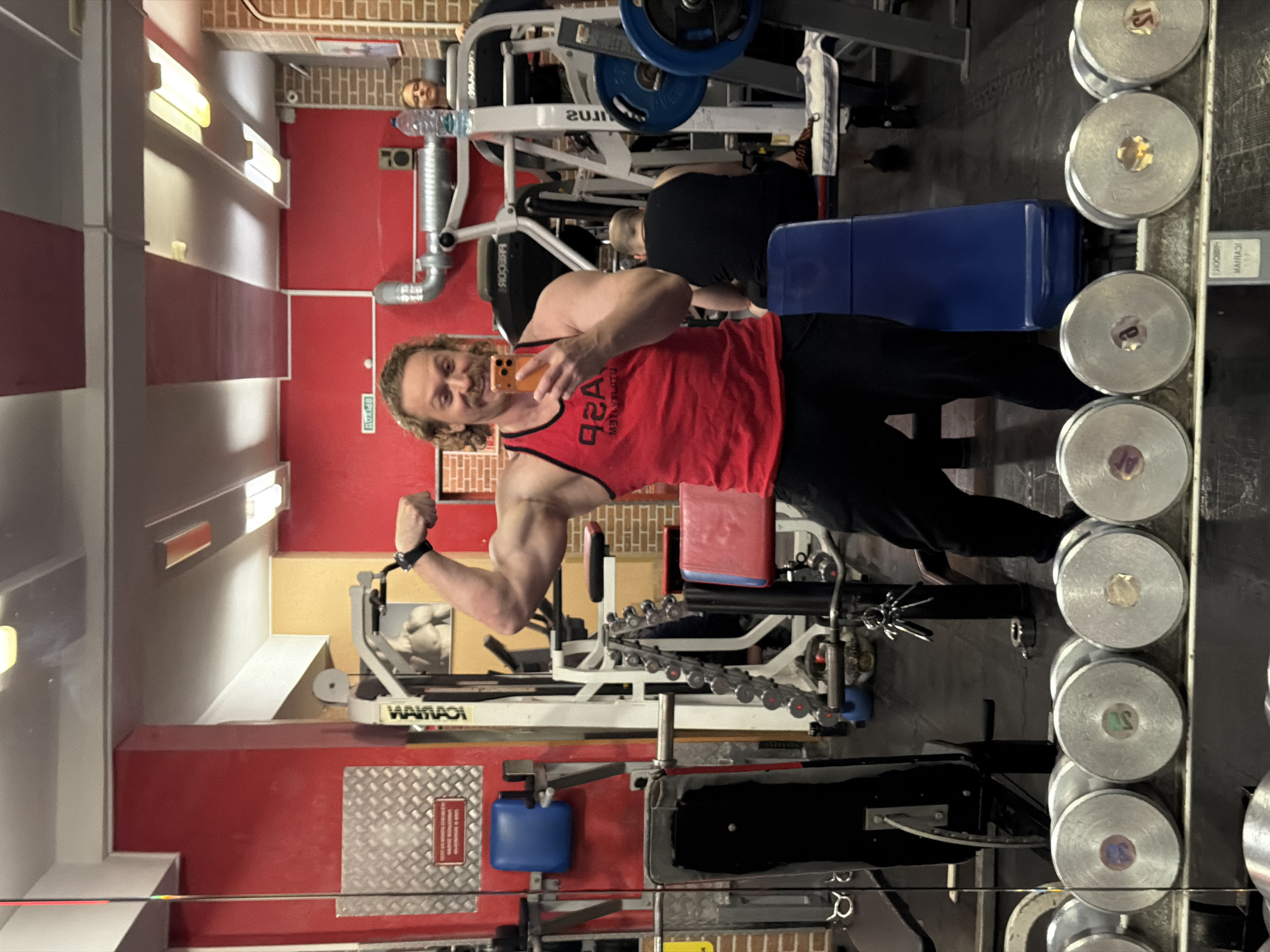 The image depicts a man in a red tank top and black pants posing in what appears to be a gym or fitness center. He is holding a cell phone and flexing his bicep. The gym has various weight machines, dumbbells, and workout equipment visible. In the background, there are other individuals using the gym. The image does not contain sufficient information to confirm the location of the gym in Saint Petersburg, Russia.