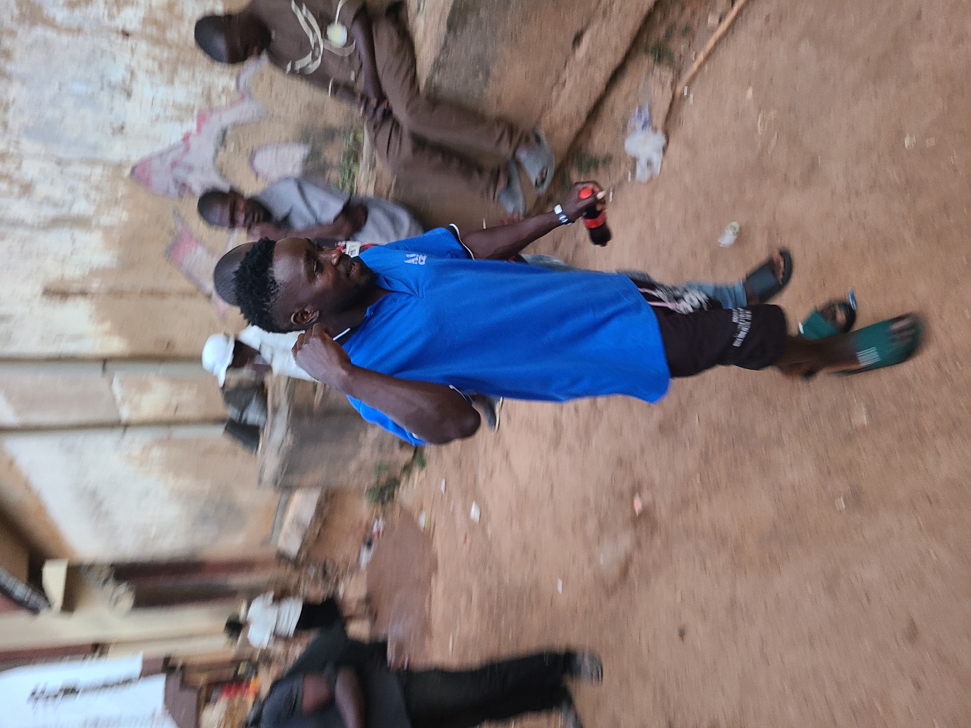 The image shows an outdoor scene in what appears to be an urban area in Jos, Nigeria. Several people are present. One man, wearing a blue shirt and black shorts, stands out prominently in the foreground and is holding what seems to be a bottle. Other people are seen lounging or sitting on the ground and on or against concrete structures in the background. The environment features sandy or dusty ground, concrete structures or walls, and scattered debris.