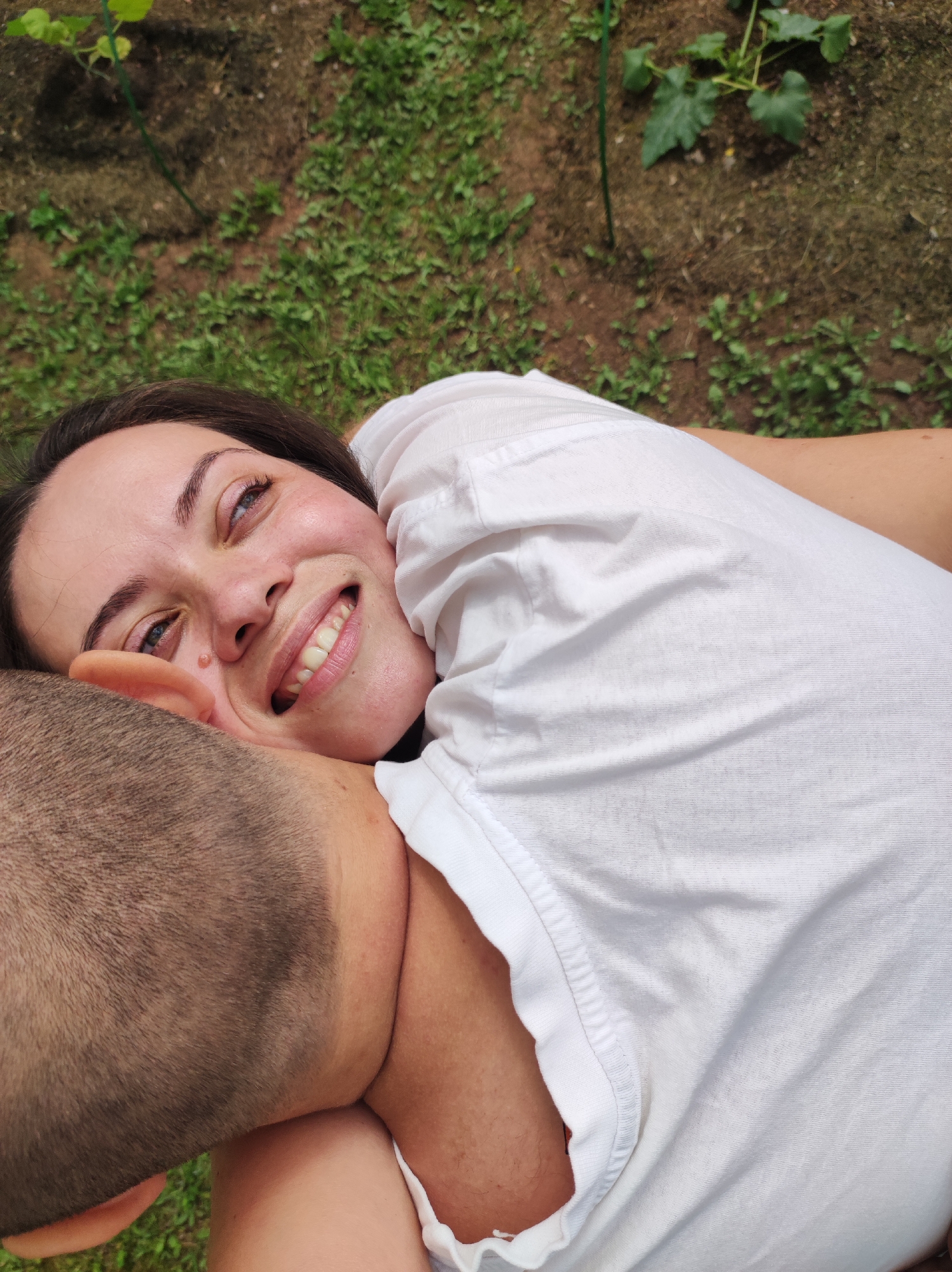This image captures two individuals, a female and a male, engaged in an intimate embrace outdoors in Foki, Russia.
The female is positioned centrally and diagonally across the lower-left to mid-right of the frame. She has dark brown hair, parted to her left, and a fair complexion with rosy cheeks. Her blue eyes are cast upwards and to the viewer's left, and she displays a broad smile, revealing her upper teeth. A small, raised epidermal lesion is visible on her left cheekbone, below her eye. She is wearing a white, short-sleeved t-shirt, which appears slightly rumpled. Her head is resting against the male subject's neck/shoulder, and her right arm is bent, with the elbow visible in the lower-left quadrant.
The male subject is partially visible on the left side of the frame, primarily his head and upper neck. He has closely cropped, dark brown hair. His back is facing the camera, and his head provides support for the female subject, indicating a prone or reclining position for both.
The subjects are situated on a surface composed of mixed green grass and exposed brown earth. In the upper two-thirds of the frame, the ground transitions to predominantly disturbed soil with sparse green vegetation. A developing plant with broad green leaves, possibly a cucurbit, is visible in the upper-center, supported by a thin green stake. The lighting suggests natural daylight conditions. The overall scene depicts a relaxed and affectionate outdoor interaction.