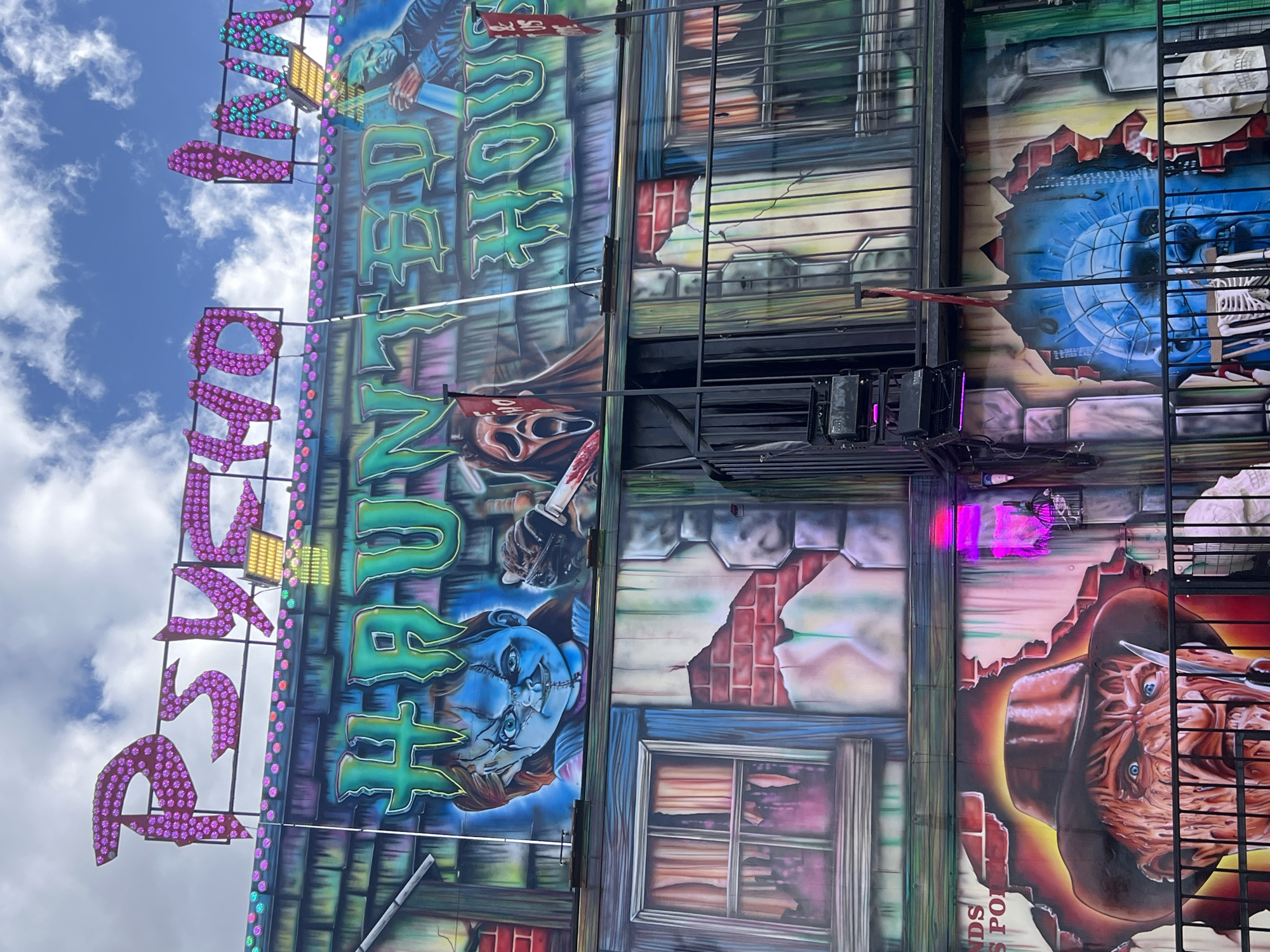 The image shows a section of a "Psycho Haunted House" ride, likely at a fair or carnival in South Norfolk, United Kingdom. The ride is decorated with images of horror movie characters, including Freddy Krueger, Chucky, and Ghostface. The words "Psycho Haunted" are displayed in large, illuminated letters. The sky is visible in the background, with scattered clouds. The ride is constructed of metal framework and painted panels.