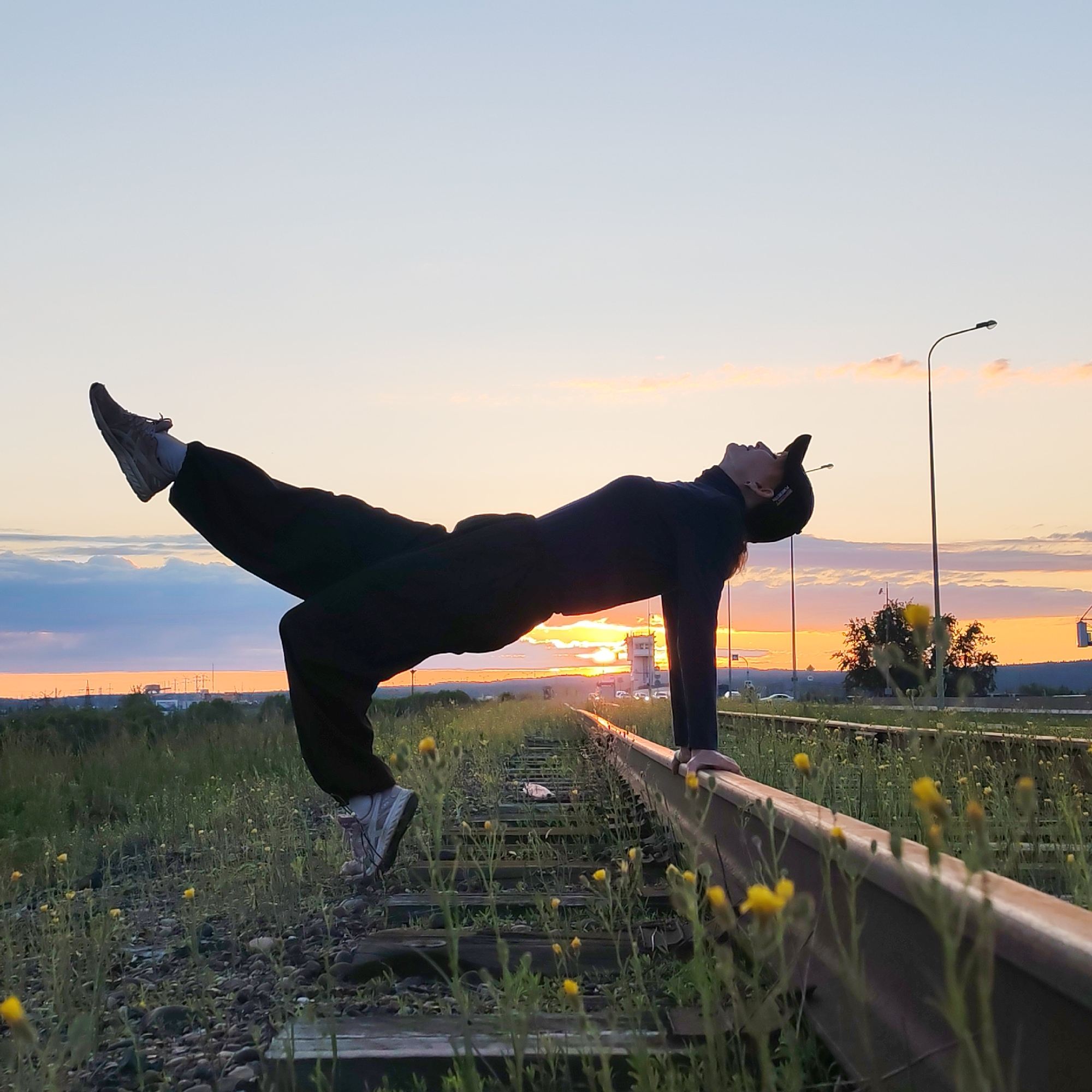 A person is captured in a dynamic, artistic pose on overgrown railroad tracks at sunset in Chaikovsky, Russia. The individual, dressed in dark long-sleeved clothing, black pants, and light-colored sneakers with white socks, performs a challenging balance or levitation-like pose. They are supported by their hands on one rail, with their head tilted back and one leg extended upwards and slightly bent. A dark baseball cap covers their head.
The setting is an outdoor, semi-rural landscape bathed in the warm light of sunset. The sky displays a vibrant gradient of orange, yellow, and pink hues near the horizon, fading into a soft blue overhead, with scattered clouds reflecting the sun's glow. The bright orange disc of the sun is visible directly behind the person's torso.
The railroad tracks, consisting of metal rails on wooden sleepers, are heavily overgrown with green grass and numerous small yellow flowers. They recede into the distance, appearing to curve slightly. On the right side of the image, a tall, dark street light with a curved arm stands prominently against the sky. In the far distance, faint industrial-like structures, possibly a control tower or factory buildings, and the silhouettes of vehicles on a road are visible on the horizon. No legible text is visible on the person or in the scene.