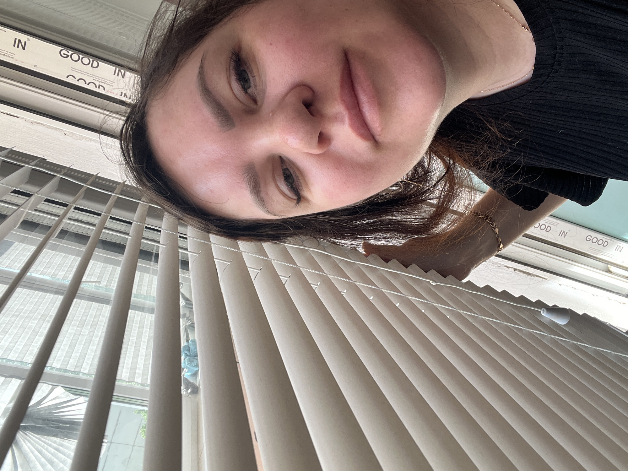 A person with brown hair and fair skin is viewed from an extreme low angle against a background of window blinds. The person is wearing a black top and a gold bracelet is visible on their wrist. The blinds are partially open, revealing a window with the words "GOOD IN" printed on the frame. The precise location in Noviy, Russia, cannot be determined from the visual information provided.