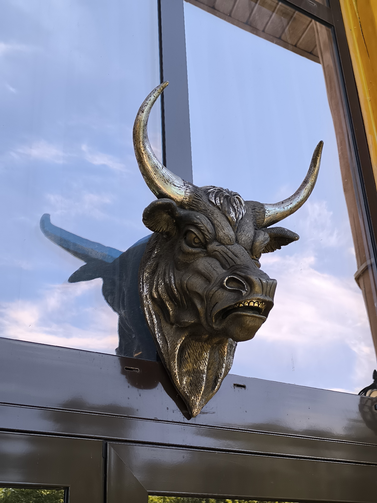 The image shows a close-up, medium shot of a bronze bull's head sculpture mounted on a window. The sculpture is dark gray with golden horns and teeth, and it appears to be emerging from the window frame, giving the illusion of it breaking through the glass. The bull has a menacing expression with its mouth open and teeth bared, and its eyes are wide and focused. The background is a reflection of the sky with scattered clouds, indicating it is likely daytime. The window is dark-colored and has a glossy surface, reflecting some of the surrounding environment. To the right of the bull's head, a sliver of a yellow structure is visible, possibly part of the building's facade. The bottom of the image shows the dark frame of the window, and a hint of greenery is reflected in the lower panes, suggesting the presence of trees or other vegetation outside. The sculpture is located on a window of a building in the city of Malyy Baklaniy, Russia.
