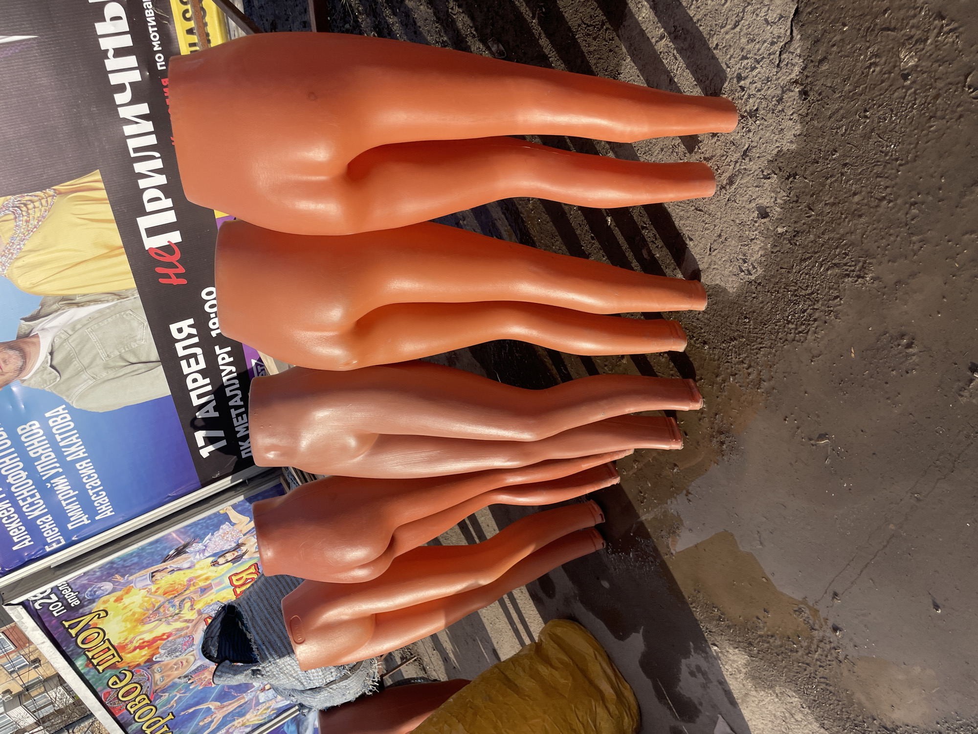 The image shows a collection of disembodied mannequin legs, of a fleshy orange color, arranged on what appears to be a street or sidewalk. The legs are stacked and oriented in the same direction. Alongside the mannequin parts are posters with Russian text and imagery. One poster advertises a show or event titled "провое шоу" and lists names such as Елена Ксенофонтов, Дмитрий Ульянов, and Анастасия Акатова. Another poster references "17 АПРЕЛЯ" (April 17th) and mentions "ПК МЕТАЛЛУРГ 10:00", suggesting a location and time. The phrase "НЕПРИЛИЧНЬ" is prominently featured on this poster. A location, most likely Izhevsk, Russia, is indicated by Cyrillic text. The environment appears to be urban, as suggested by the sidewalk and the posters advertising local events. The lighting suggests daylight. The surface where the legs are placed appears to be somewhat dirty or stained.