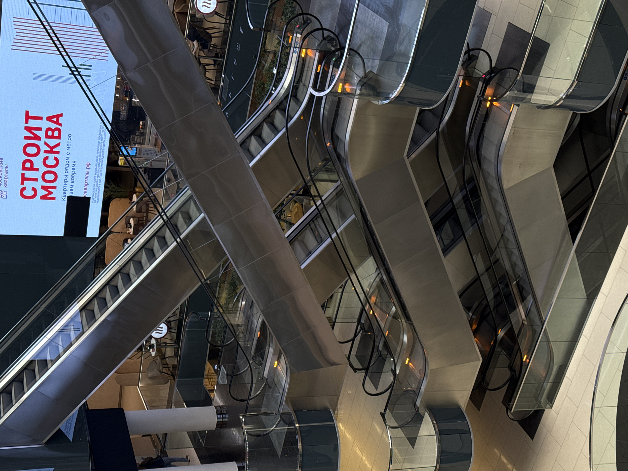 This indoor shot depicts a building interior in Khimki, Russia. Multiple escalators dominate the scene, ascending and descending at different angles. A large screen displays an advertisement in Russian, "СТРОИТ МОСКВА," related to apartments near the metro, along with the website "скварталы.рф." The environment seems modern, with neutral tones and lighting. The overall impression is of a bustling urban space, likely a shopping mall or transport hub. No people are clearly visible.
