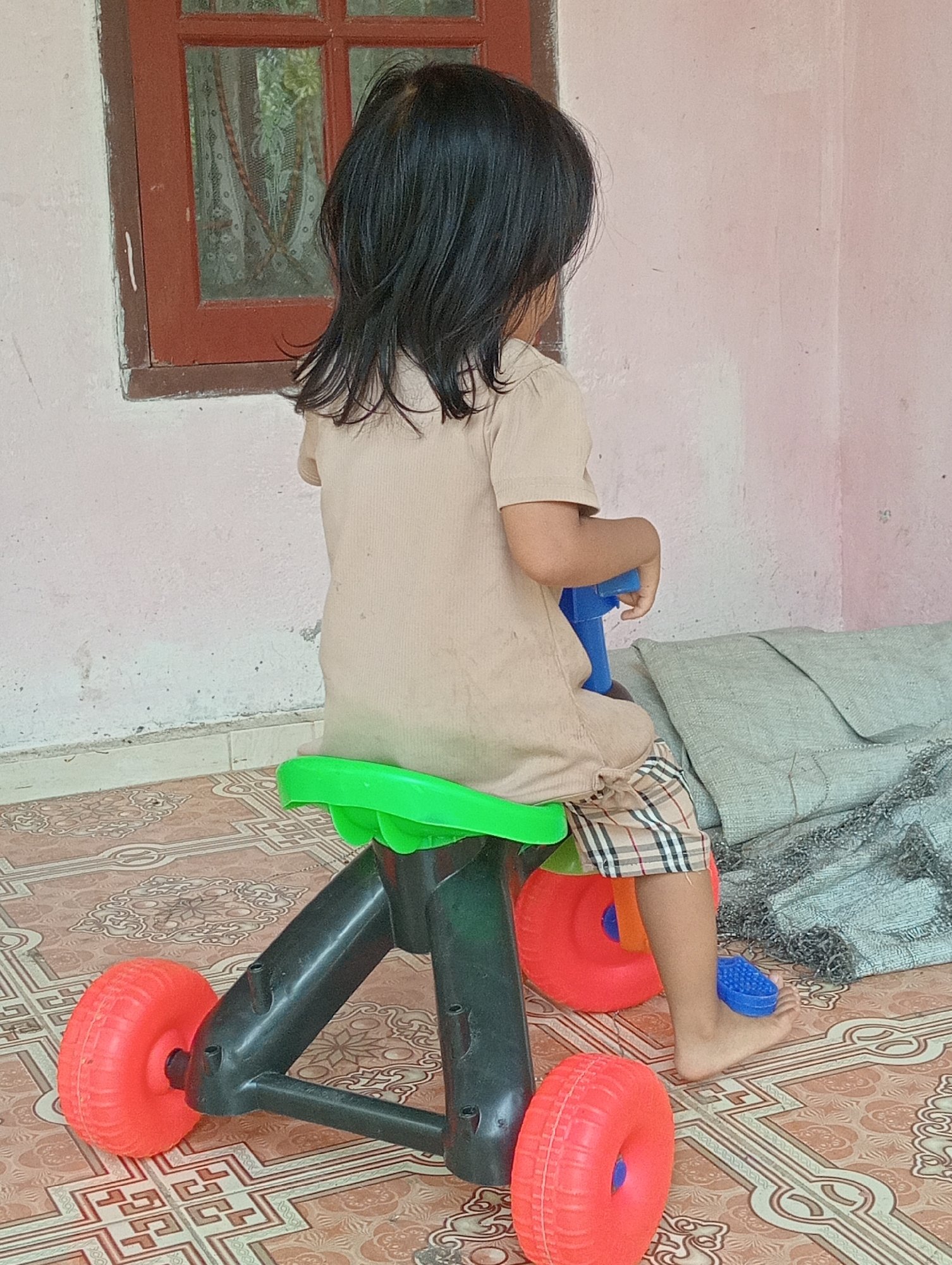 A young child, viewed from the back, sits on a green plastic toddler tricycle with orange wheels. The child has long, dark hair and is wearing a light tan short-sleeved shirt and plaid shorts. Their right foot, clad in a blue plastic shoe, rests on a blue pedal attached to the tricycle. The tricycle is positioned on a tiled floor with a patterned design. In the background, a light pink wall is visible, with a red-framed window on the left. To the right of the tricycle, a folded gray blanket or fabric is placed on the floor. The scene appears to be outdoors or in a covered outdoor area, potentially a porch or patio, located in Campago Barat, Indonesia.