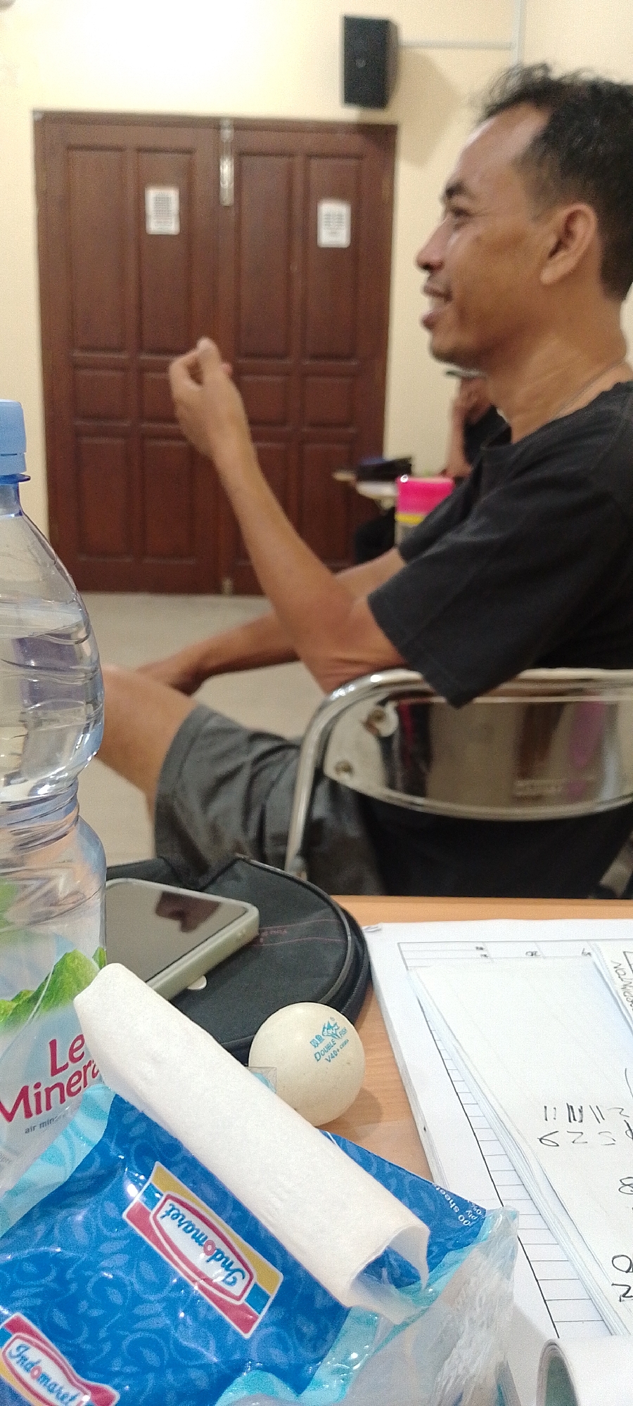 A man in a black t-shirt and grey shorts is seated in a metal chair in what appears to be an indoor room. He is facing to the right and looking upwards. His right hand is raised and appears to be holding something small. In the foreground, there is a plastic water bottle, a smartphone, a white cylindrical object, a ping pong ball, and a blue package with Indonesian text. Behind the man, there are double wooden doors with a light-colored wall and a black speaker mounted high on the wall. The environment suggests a casual setting, possibly a common room or a classroom in Kudus, Indonesia, given the presence of the Indonesian text on the packaging.