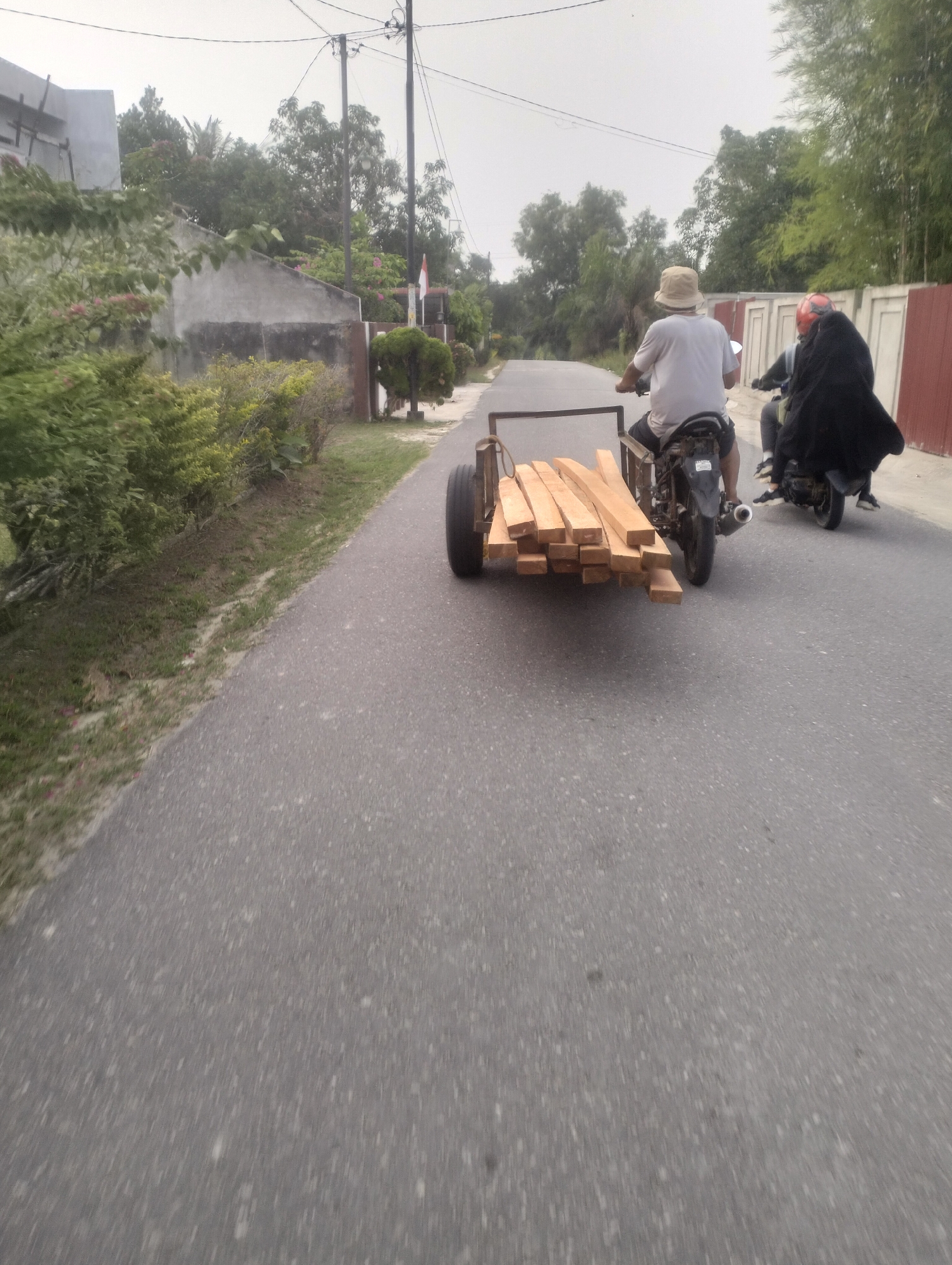 In Simpang Padang, Indonesia, a man wearing a light-colored bucket hat and a white t-shirt rides a motorcycle that is towing a small trailer laden with lumber. The trailer has a single black wheel on the right side. Behind the man, another motorcycle is visible, carrying a passenger dressed in black with a red helmet. Both motorcycles are traveling down a paved road lined with vegetation on the left and a fence with red panels and concrete posts on the right. The environment suggests a semi-rural or suburban setting with scattered trees and a building visible in the background. The sky is overcast.