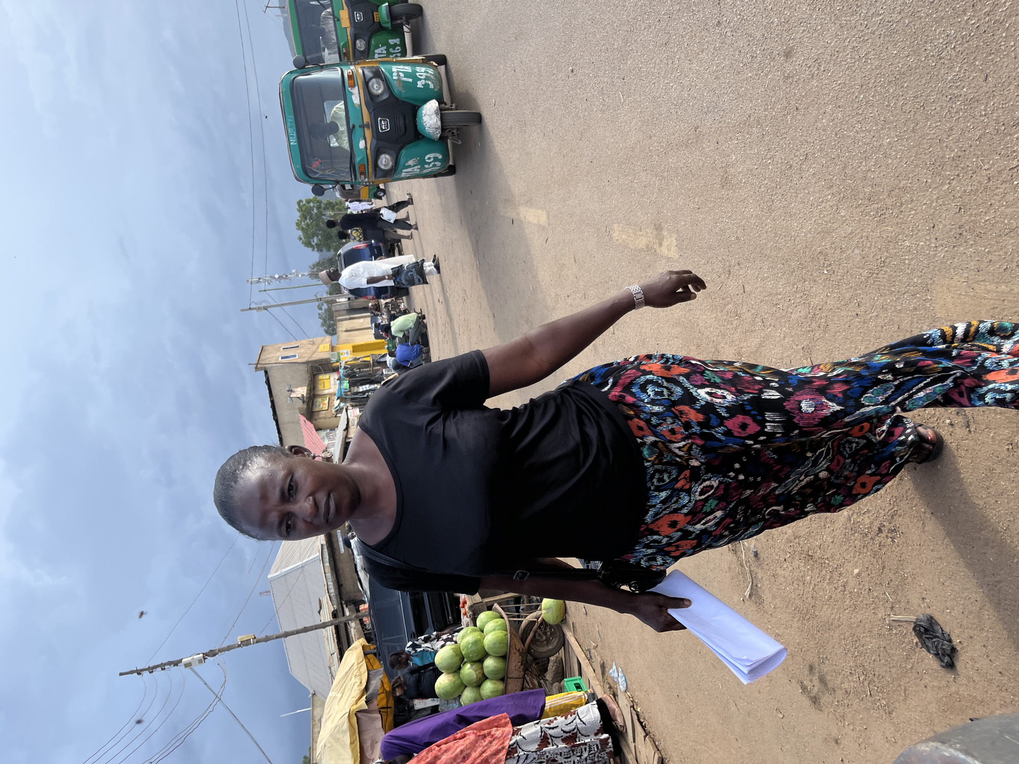An overcast day in Rafin Par, Nigeria, shows a woman in a black t-shirt and a colorful, patterned skirt walking on a dirt road. She carries a rolled-up document in her left hand and wears a watch on her left wrist. In the background, several auto-rickshaws, some green and some yellow, are parked or driving on the road. To the left of the woman, a cart piled with green fruits, possibly watermelons, is visible, with market stalls and buildings lining the street. Other people, including a man in a white outfit, are scattered throughout the scene, engaged in various activities. The environment appears to be a bustling marketplace with a mix of urban and semi-rural elements.