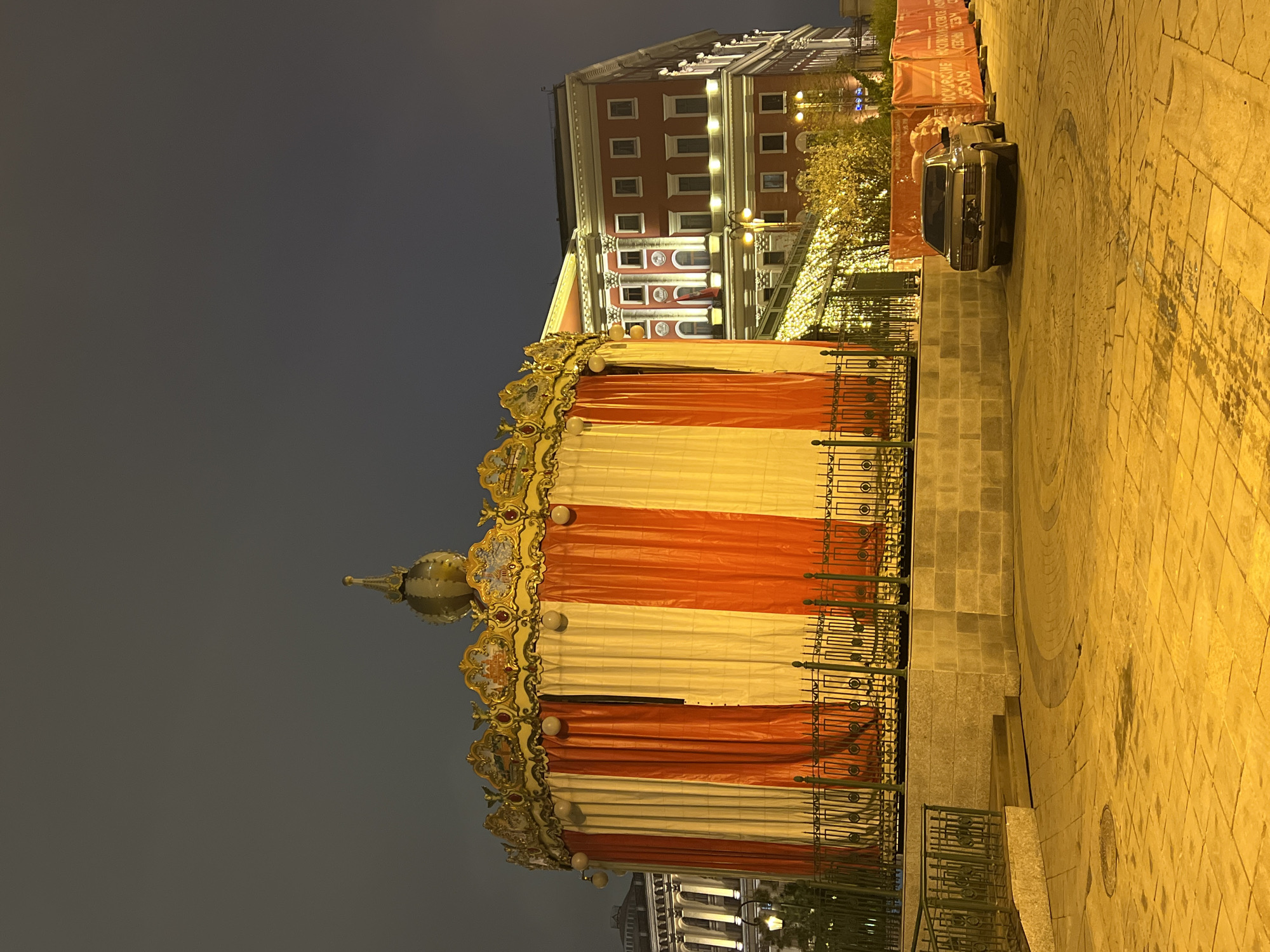 The image shows a carousel in Moscow, Russia at night. The carousel is closed and draped with red and white striped material. The carousel is ornate with a golden crown and globe on top. A black car is parked to the right of the carousel. A large building is in the background. The area is lit by streetlights.