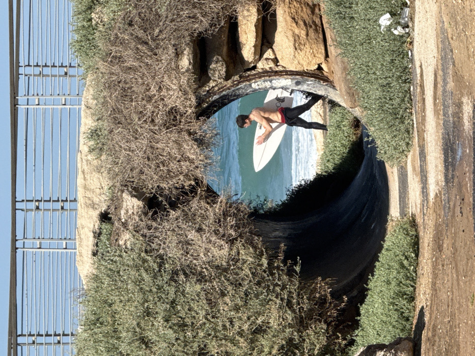 The image displays a coastal scene in Tel-Aviv, Israel, centered on a natural-looking underpass leading to a beach.
**Environment:** The foreground features a dirt path with patches of low green vegetation, leading into a dark, semi-circular underpass. This underpass is constructed with large, light-colored rocks and is heavily overgrown with dense green bushes on the left and dry, brown, tangled scrub on the right, creating a tunneled effect. Above the underpass, a modern metal railing, characterized by vertical posts and multiple horizontal bars, is visible against a clear blue sky, suggesting an elevated promenade or pathway. Through the underpass opening, a light-colored sandy beach is visible, meeting the turquoise Mediterranean Sea, which exhibits white breaking waves. On the ground in the lower right, outside the tunnel, discarded waste including plastic bottles and a white bag is present.
**People:** One individual, a young male, is seen on the sandy beach, positioned to the right of the underpass's exit. He is shirtless, wearing dark long pants, possibly a wetsuit bottom or athletic wear, with a red waistband visible. He has dark hair and a lean physique.
**Actions and Objects:** The male subject is walking towards the right side of the frame, carrying a white, shortboard-style surfboard vertically against his right side, holding it with both hands. His left foot is forward, indicating movement along the beach.