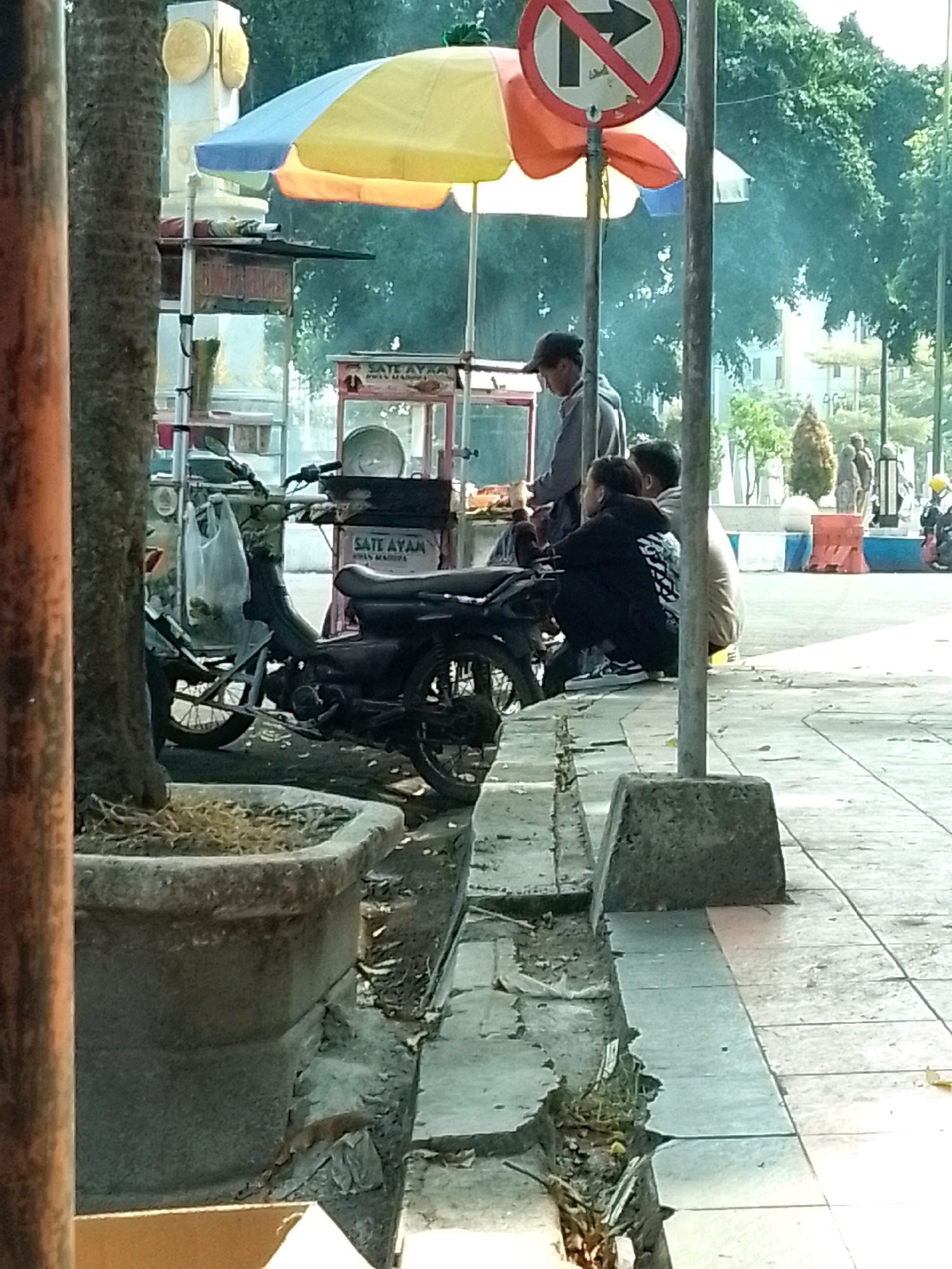 The image depicts a street scene in Jember, Indonesia, likely a public area with pedestrian pathways. In the foreground, a large concrete planter with dry vegetation is visible on the left. A wooden pole is positioned in front of the planter. Adjacent to this, a motorbike is parked, with its front wheel facing towards the right of the frame. A food cart is set up behind the motorbike, featuring a yellow and orange umbrella. Text on the cart reads "SATE AYAM".
To the right of the food cart, three individuals are present. One person, wearing a dark baseball cap and a light-colored hooded jacket, appears to be interacting with the food cart. Two other individuals are seated on the ground, facing the food cart. One is wearing a black hooded jacket with white lettering on the back, and the other is wearing a light brown jacket.
In the background, lush green trees form a backdrop, suggesting an outdoor environment. A traffic sign with a red circle and a black arrow indicating "no right turn" is mounted on a pole to the right of the food cart. Further in the distance, buildings and more trees are visible, along with a few other people. The ground is paved with tiles, and a concrete curb runs along the pathway. The lighting suggests it is daytime, with sunlight casting shadows.