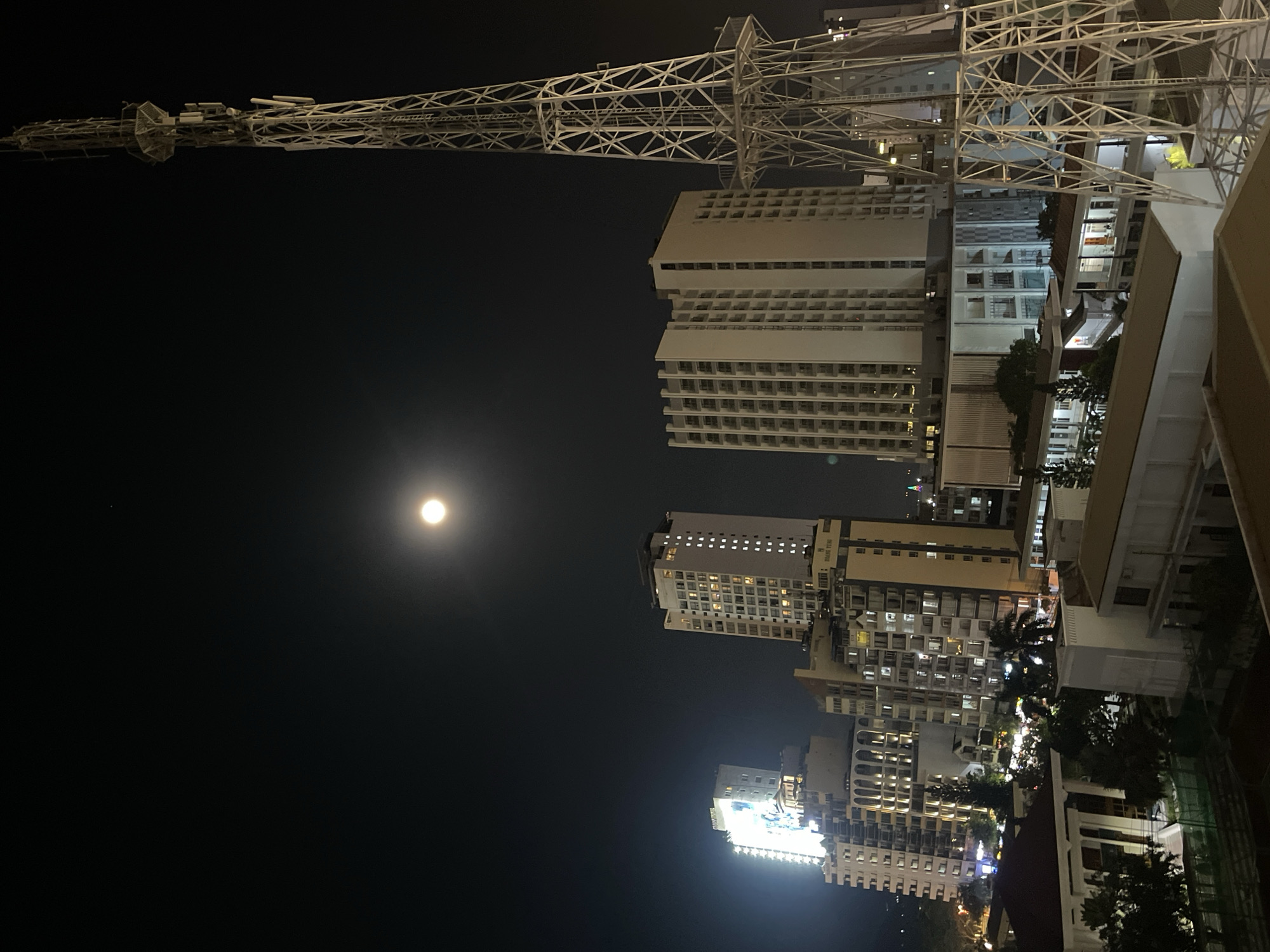 The image shows a night scene in Nha Trang Ward, Vietnam. A tall white metal tower structure dominates the upper left, extending from the upper edge. Below, a cluster of illuminated high-rise buildings rise into the night sky. A full moon is visible in the upper left, separate from the lit buildings.