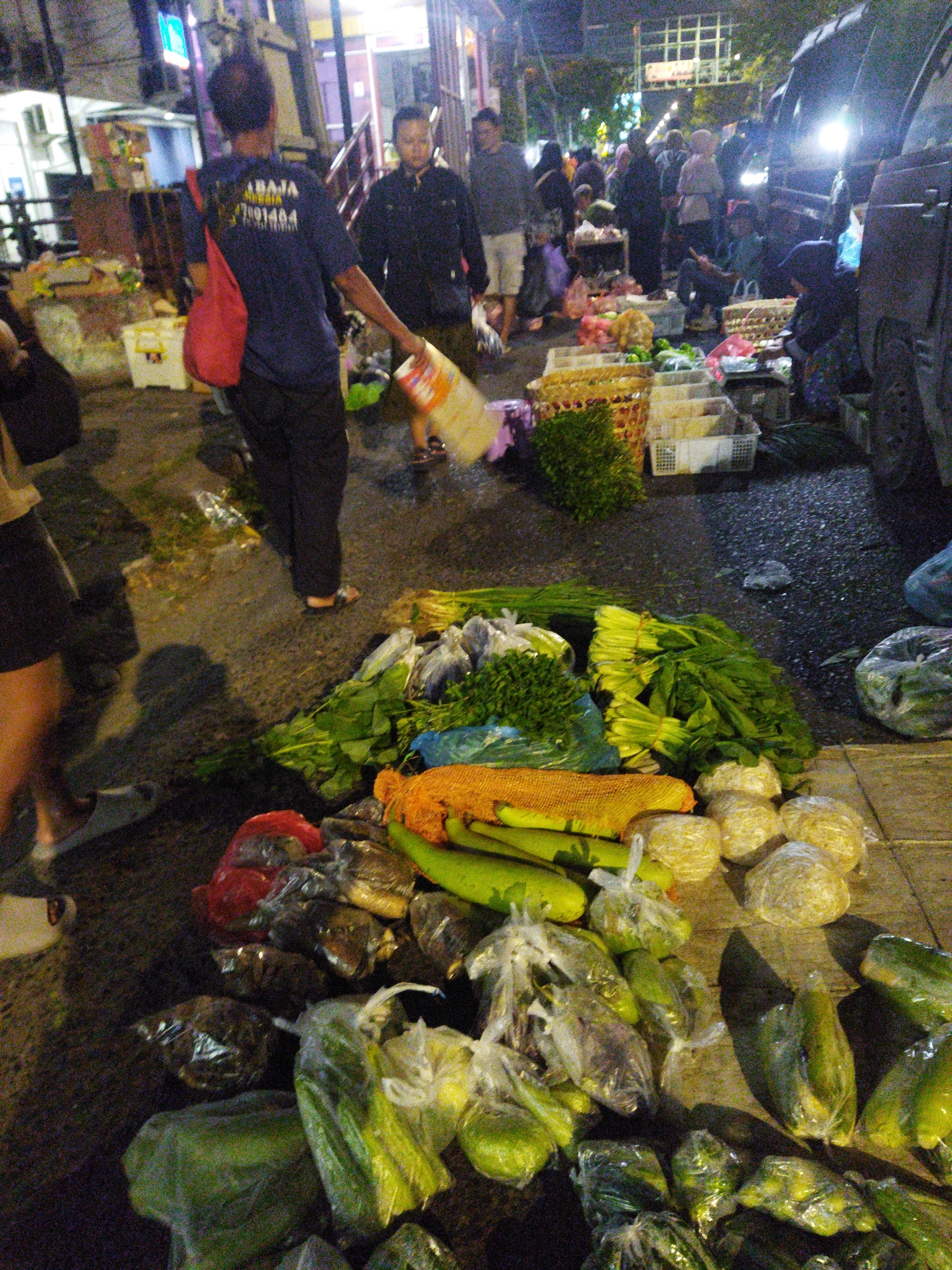 This is a nighttime street scene in Semarang, Indonesia, likely a market or roadside stall area. A man in a dark blue t-shirt with yellow lettering and a red bag slung over his shoulder is walking and carrying a large plastic bag. To his right, other people are visible, some interacting with goods on display. The ground is paved and appears wet. Piles of fresh produce, including leafy greens, cucumbers, and what appear to be root vegetables or gourds, are arranged in plastic bags and baskets, some on the ground and others on what seem to be makeshift stalls or tables. A dark-colored vehicle, possibly a van, is parked on the right side of the image, its headlights illuminating some of the scene. The lighting suggests it is after dark, with artificial lights from nearby structures and the vehicle contributing to the illumination.