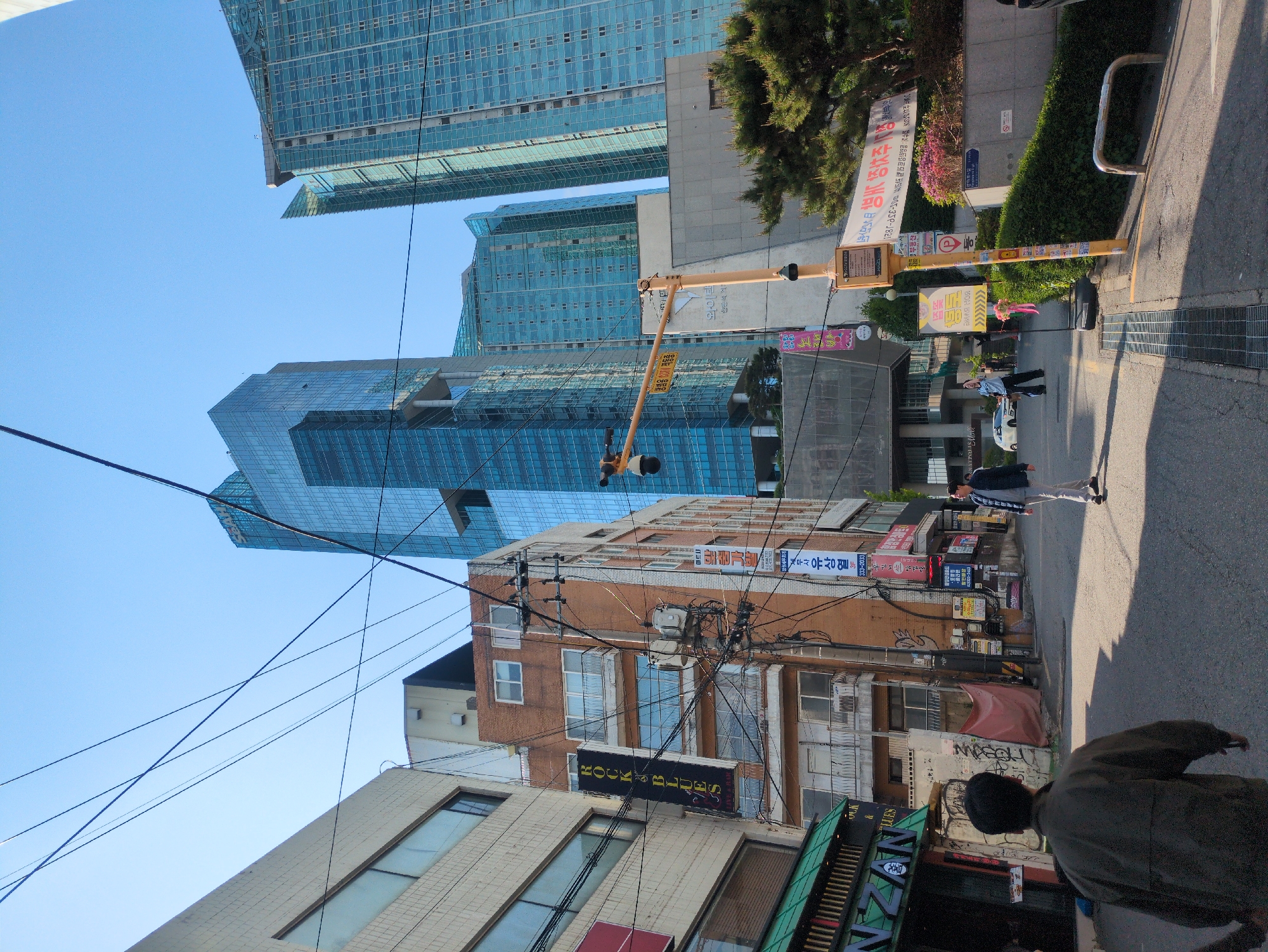 The scene captures an urban street in Seoul, South Korea, presenting a mix of modern skyscrapers and older buildings. Three tall, blue-tinted glass skyscrapers dominate the upper section of the image, contrasting with the lower part that shows older, brick and concrete buildings lined along the street. Electrical wires crisscross in the sky, adding to the urban density.
On the right side of the image, people are walking along the sidewalk, indicating street activity. Signs in Korean are visible on the buildings, including one mentioning "ROCKS BLUES". Graffiti and other street art are present on the lower buildings. A person in a dark jacket is partially visible in the foreground. The composition suggests a blend of old and new architecture, typical of Seoul. The lighting suggests it is daytime.