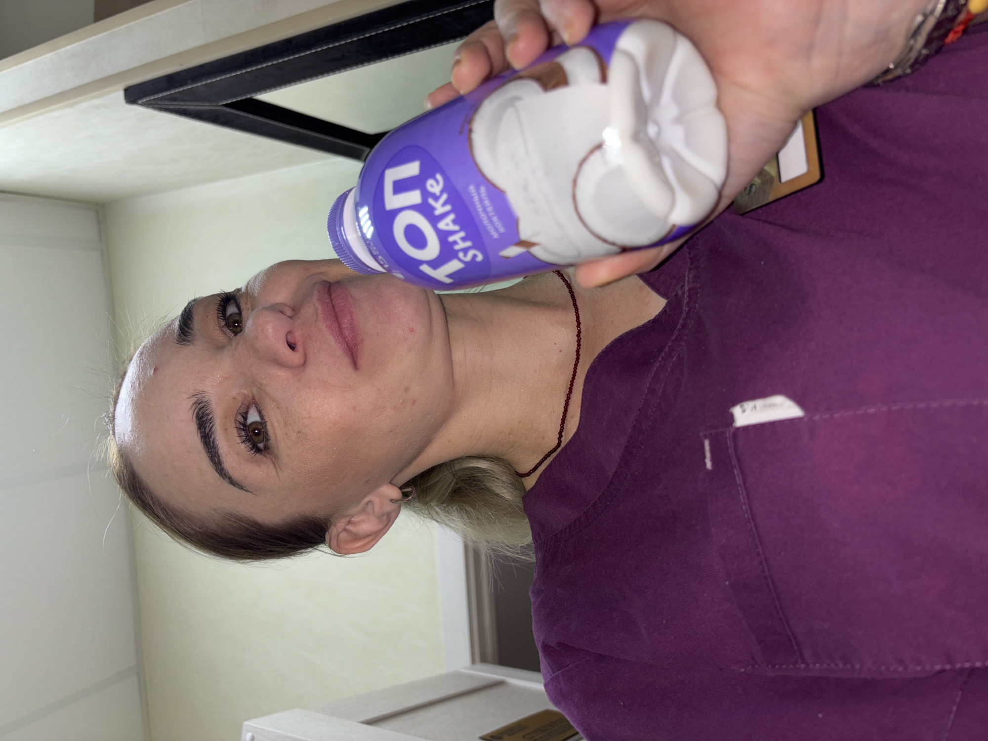 A woman in a purple scrub top is holding a bottle of "TON SHAKE" flavored with coconut. The bottle is purple and white and shows an image of coconut. The woman's face is partially visible, and she has on makeup. The background appears to be indoors, possibly a kitchen or bathroom, with light-colored walls and a dark-framed mirror above a counter. The woman has blond hair and is wearing a thin, beaded necklace. The image is oriented sideways.