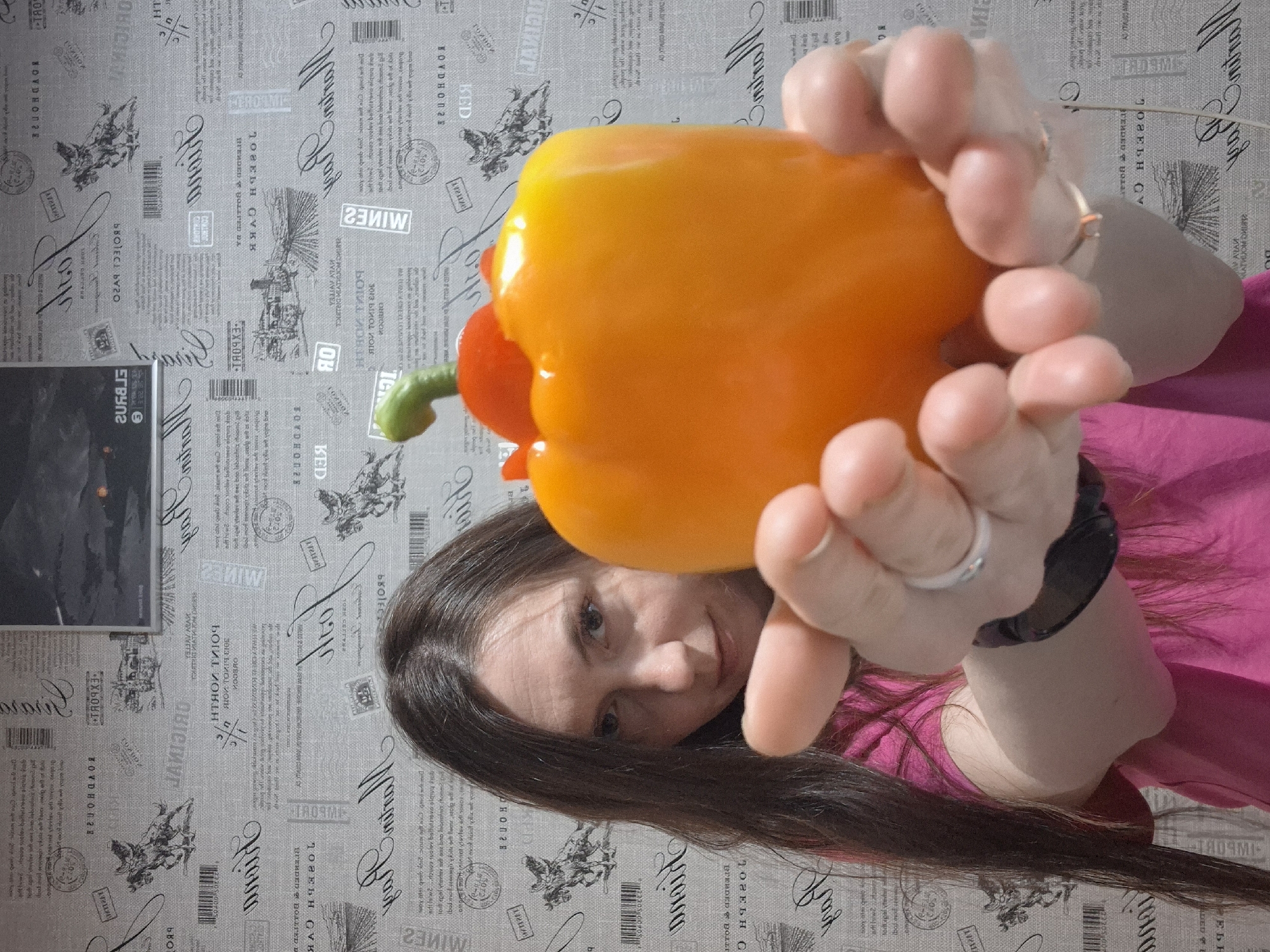 A woman is holding an orange bell pepper in front of her face. She is wearing a pink shirt and a black watch. Her long brown hair is partially obscuring her face. The background is a gray wallpaper with vintage-style text and illustrations. There is a dark rectangular object hanging on the wall behind her.