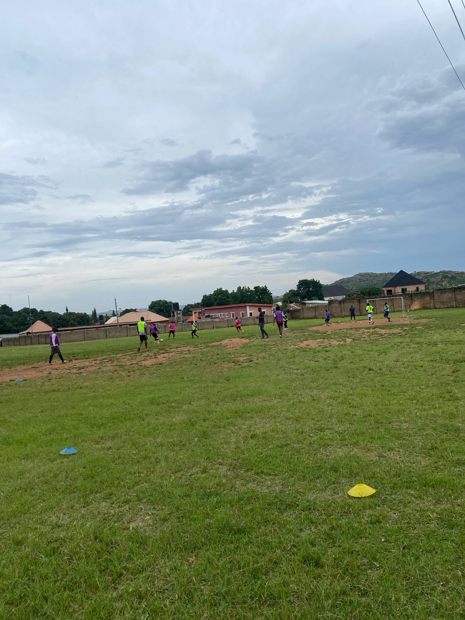 This image depicts an outdoor football training session taking place on a grassy field. Several individuals, dressed in athletic wear, are actively engaged in the sport, with some interacting with a soccer ball. The background reveals a residential area with buildings and greenery, suggesting an urban or suburban environment. The sky is overcast with clouds. There are two colored cones visible on the grass, one blue and one yellow, which are likely used for drills. The scene appears to be located in Jos, Nigeria, given the architectural styles of the buildings and the overall landscape.