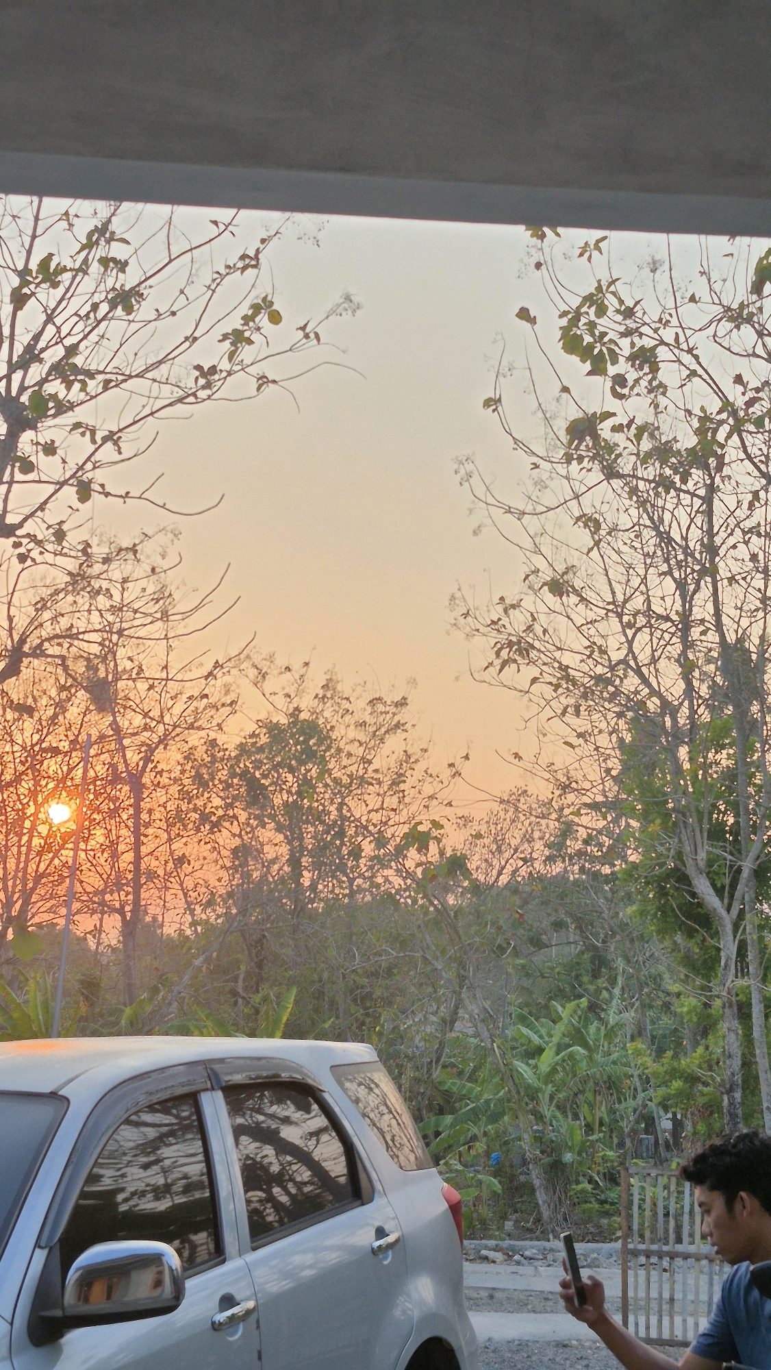 A silver SUV is parked on a gravel surface near a wooden fence. The car's windows reflect the surrounding trees and the orange sky. A young man is seated on a motorcycle to the right of the car, looking at his phone. The scene is set against a backdrop of trees and vegetation under a warm, overcast sky that suggests either sunrise or sunset. The location is identified as Gambiranom, Indonesia.
