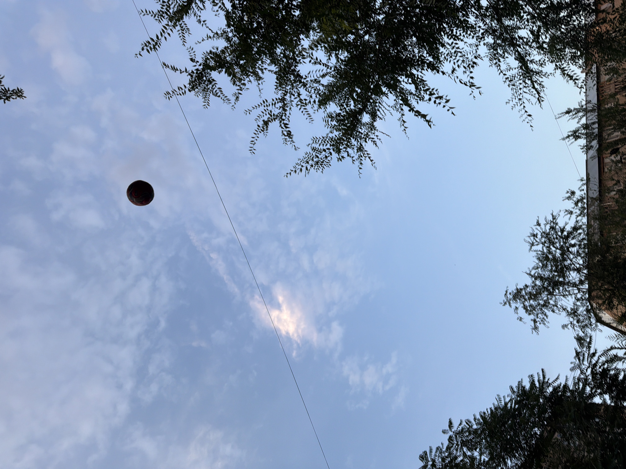 This image is an outdoor shot of the sky, looking upwards. The sky is a light blue with scattered white clouds. Sunlight is visible breaking through a cloud in the upper right quadrant.
In the left-center of the frame, a dark, spherical object, possibly a ball or a decorative lantern, is suspended in the air. It has a visible pattern of red and black shapes. A thin, dark line, likely a wire or cable, extends diagonally from the upper right corner of the image towards the center, passing near the suspended object.
The foreground is partially obscured by dark green foliage from trees or bushes in the upper and lower parts of the frame, and a portion of a brick building is visible on the far right edge. The foliage and the building suggest an urban or semi-urban environment.
The lighting and the light blue color of the sky indicate it is daytime, likely late afternoon or early evening given the soft sunlight. The presence of clouds suggests it is not a perfectly clear day. There is no visible text in the image. The overall mood is calm and perhaps a little serene, with the suspended object adding a touch of intrigue.