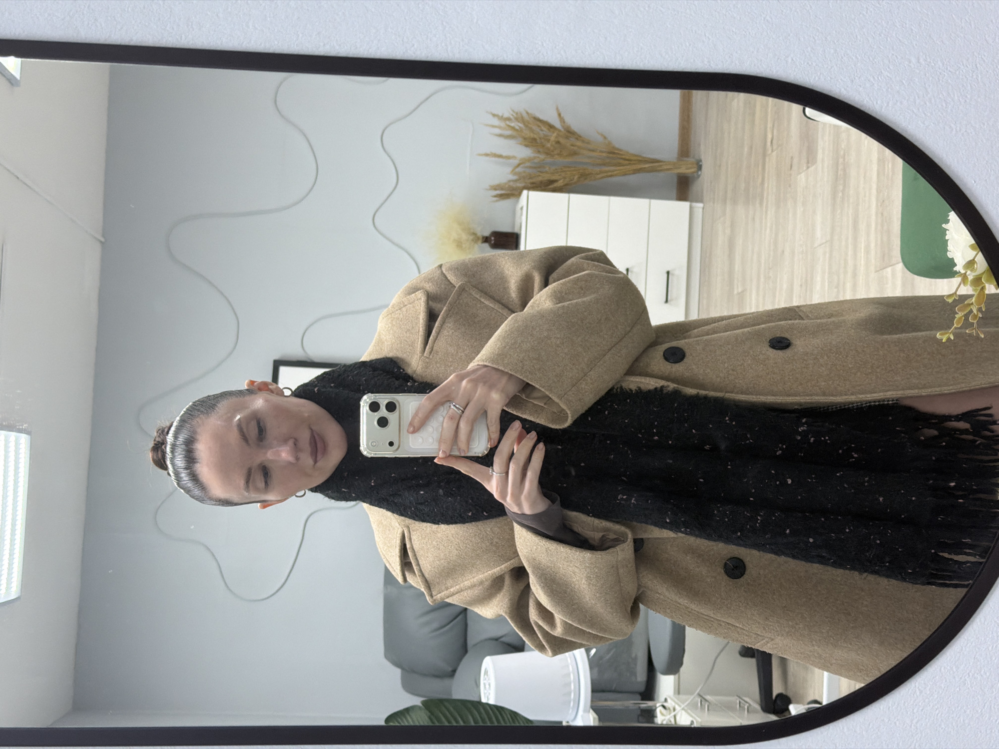 A woman takes a selfie in a mirror. She is wearing a tan coat and a dark scarf. The mirror reflects a room with a gray couch, a white dresser with wheat stalks on top, and a black-framed picture on the wall. Based on the user’s location, this is likely occurring somewhere in the city of Perm, Russia.