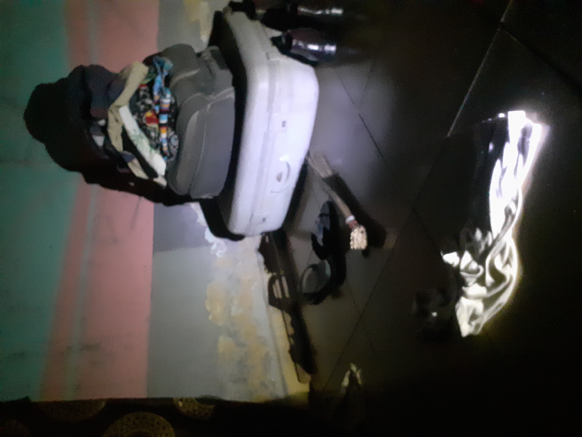 The image depicts a dimly lit room, likely in Jalingo, Nigeria, cluttered with various items. In the foreground, a pile of clothes, some colorful and patterned, is stacked on top of a dark suitcase, which in turn rests on a white rectangular object, possibly another suitcase or a storage container. Several pairs of flip-flops are scattered on the dark tiled floor. A white cloth is draped over the floor in the lower left portion of the image, reflecting light. In the background, a wall features a mural or painted design with horizontal bands of blue, pink, and beige. The overall impression is one of disarray and an unorganized living space.