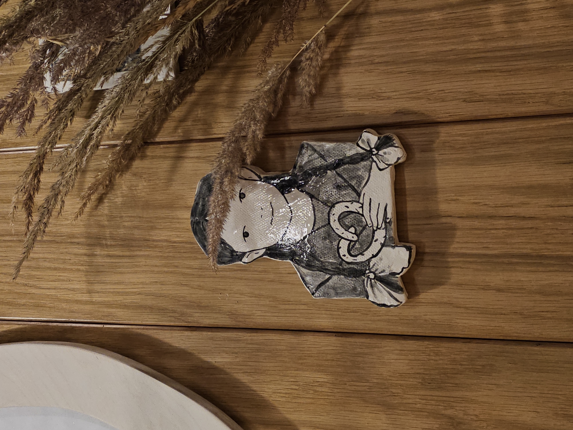 A ceramic cutout of a young girl with braided hair, wearing a checkered top, and holding a pretzel-shaped object is affixed to a wooden wall. The girl has a white face with black eyes and a simplified smile. Her hair is dark and pulled into two braids tied with ribbons at the ends. The cutout has a slightly glossy finish. The wooden wall is made of planks with visible grain. A bunch of dried grass stalks, with feathery tops, is placed to the right of the cutout, casting shadows on the wall. A portion of a light-colored, oval-shaped object, possibly a mirror frame, is visible on the far left of the image. The scene is indoors. The location is Nemchinovo, Russia.