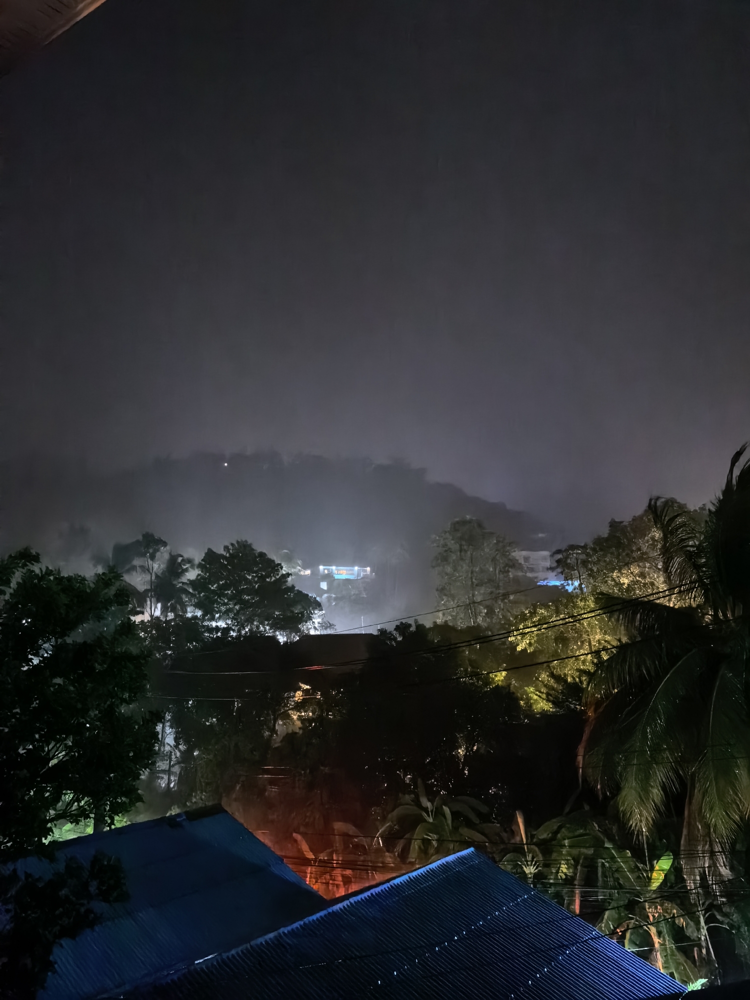 The image depicts a nighttime scene in the city of Ko Samui District, Thailand. The view is from a high vantage point, looking over a landscape characterized by dense vegetation and some buildings. The sky is dark and obscured by fog or mist, which creates a hazy, atmospheric effect, particularly over the distant hills.
In the foreground, the corrugated roofs of two buildings are visible, one a deep blue and the other a darker, less defined color. These roofs appear to be made of metal. Above and between these structures, a dense canopy of trees dominates the mid-ground. Various species of trees are present, with lush green foliage, some of which is illuminated by artificial light, creating patches of yellow and green. Palm trees are identifiable, with their distinctive fronds.
In the distance, several buildings are discernible through the mist, emitting lights, some of which are brightly colored, appearing as blues and whites. These structures are nestled within the hillsides. Power lines crisscross the scene in the mid-ground, adding a man-made element to the natural landscape.
The overall impression is one of a tropical, humid environment at night, with low visibility due to fog and darkness, punctuated by the presence of artificial lights from buildings and possibly street lamps. There are no people or overt human activity visible in the frame.