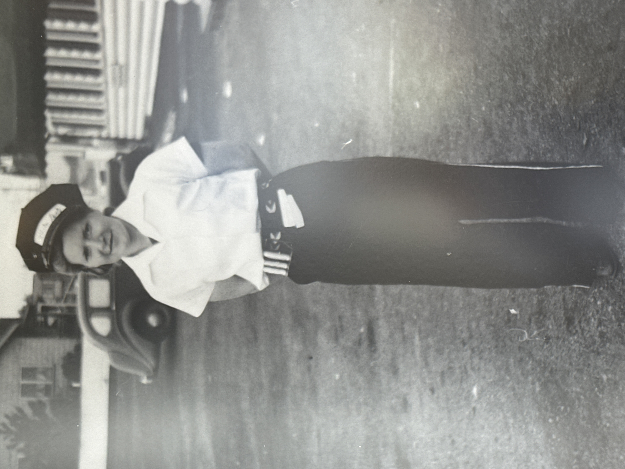 A black and white photograph depicts a woman standing outdoors in what appears to be the Township of Langley, Canada. She is wearing a uniform consisting of a white short-sleeved shirt, a dark skirt or pants, and a hat with light-colored lettering. She smiles at the camera. To her left is a car, and behind her, a building. The ground appears to be grass or dirt.