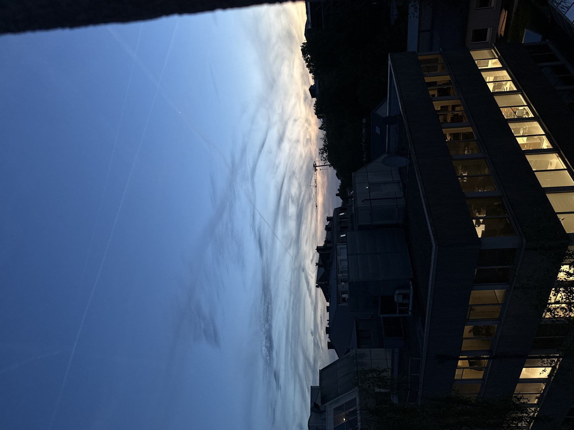 The image captures an urban or suburban evening scene, likely in Luxembourg, during dusk or early night. The sky, which occupies the upper two-thirds of the frame (when oriented correctly), displays a gradient of deep indigo blue at the zenith, softening to lighter blues and purples closer to the horizon. Streaks of wispy, elongated clouds stretch across the sky, catching the last warm orange and yellow light from the setting sun, creating a painted effect. Several faint contrails from aircraft are visible traversing the blue expanse.
The lower and right portions of the image feature several dark, modern buildings. A prominent structure on the right side of the frame showcases multiple stories with numerous rectangular windows, many of which are illuminated with warm, golden light. These lights offer glimpses into interior spaces, suggesting occupied offices or residences. Further back, a construction crane is faintly visible against the horizon line. Dark silhouettes of trees line parts of the horizon, adding to the urban landscape. The overall mood is quiet and contemplative, reflecting the peaceful transition from day to night in a developed area. No visible text is present in the image.