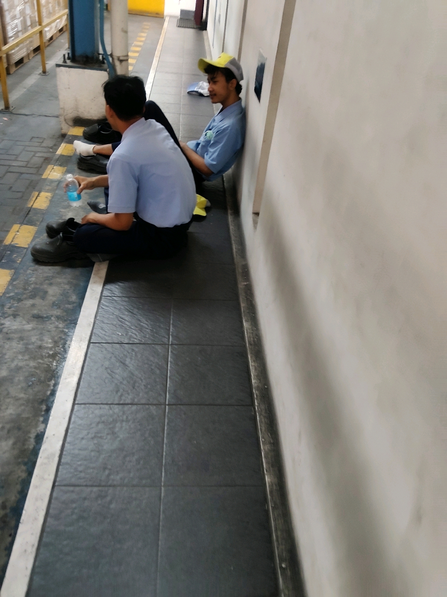 Two young males, appearing to be workers, are seated on the ground against a lightly colored wall. Both are wearing light blue collared shirts and dark trousers. The male on the left is facing away from the camera, holding a clear plastic bottle with a blue liquid. The male on the right is facing left, wearing a yellow and gray baseball cap, and appears to be looking towards the other male. The ground is a combination of grey concrete with yellow striping and dark tiled flooring. In the background, there are stacks of pallets and industrial equipment. The scene is likely an outdoor or semi-outdoor industrial area in Mekarmukti, Indonesia.