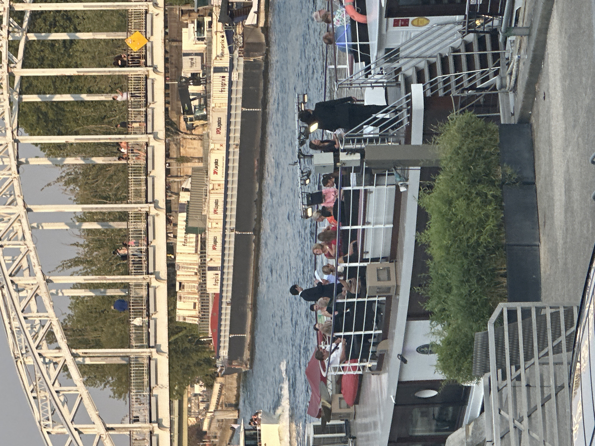 The image depicts a scenic view of the Seine River in Paris, France, with a prominent white metal bridge arching overhead. Several barges are docked along the river, some carrying shipping containers. In the foreground, a white boat is moored, with people visible on its deck. Some individuals are seated, while others stand and converse. A man in a suit appears to be addressing the group, holding a document. To the right of the boat, a set of metal stairs leads up to another level. Lush green plants are situated in a dark rectangular planter on the boat's deck. The scene is illuminated by natural light, suggesting it is daytime.