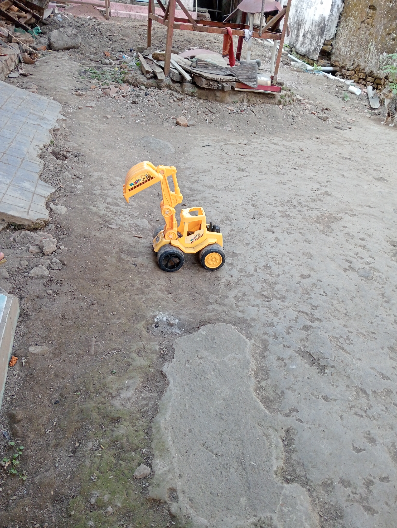 A bright yellow toy excavator with black wheels and a scoop sits on a dirt and concrete ground in what appears to be an outdoor, possibly rural, area in Desa Silo, Indonesia. The ground is uneven, with patches of dry earth, some concrete slabs, and scattered debris. In the background, there is a structure with wooden supports and corrugated metal, suggesting a construction or storage area. A small tabby cat is visible in the upper right portion of the image, near a weathered wall. The scene is illuminated by natural light.