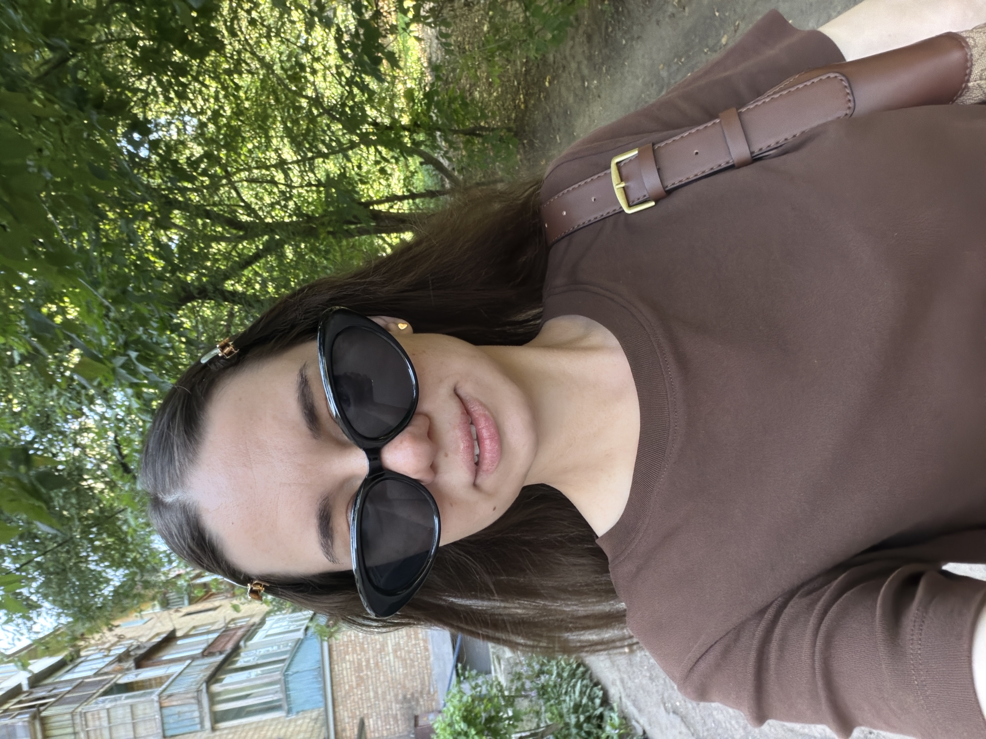 A woman is taking a selfie outdoors, in what appears to be an urban environment with trees and a building. She is wearing black cat-eye sunglasses, a brown t-shirt, and a brown crossbody bag with a gold buckle. Two hair clips can be seen in her brown hair. Behind her, on the left, is a multi-story building with balconies, constructed of brick and painted sections, and on the right, a path lined with trees and foliage. The lighting suggests it is daytime. The location is identified as Kyiv, Ukraine.
