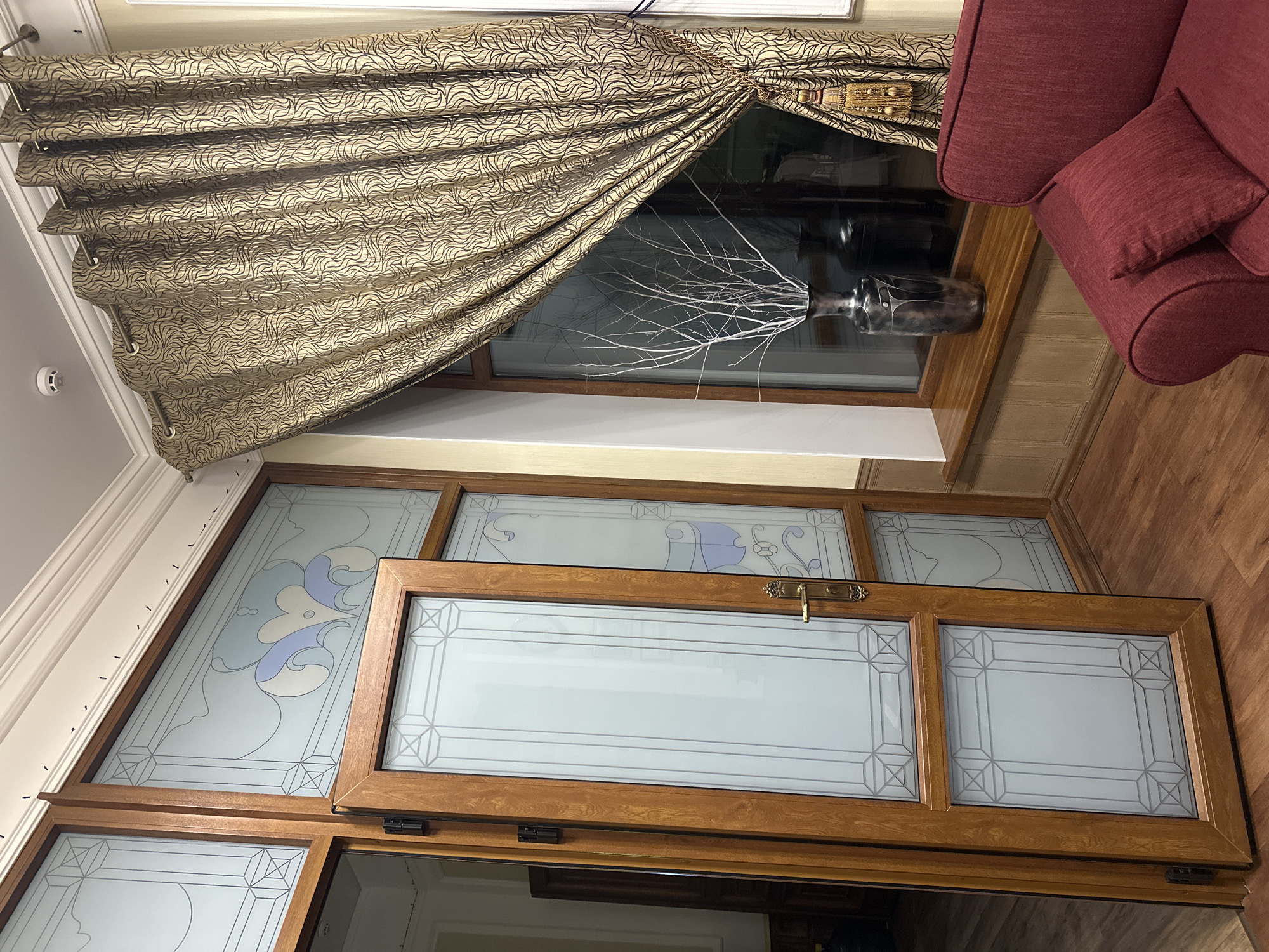 The image shows an interior scene with wooden framed doors featuring frosted glass panels with decorative lines. To the right, a patterned curtain hangs over a window, partially obscuring a dark vase containing white branches. A portion of a red sofa with a throw pillow is visible in the lower right corner. The scene appears to be a room in a building. Without further information or context, it is impossible to determine the specific location within Khvalynsk, Russia.
