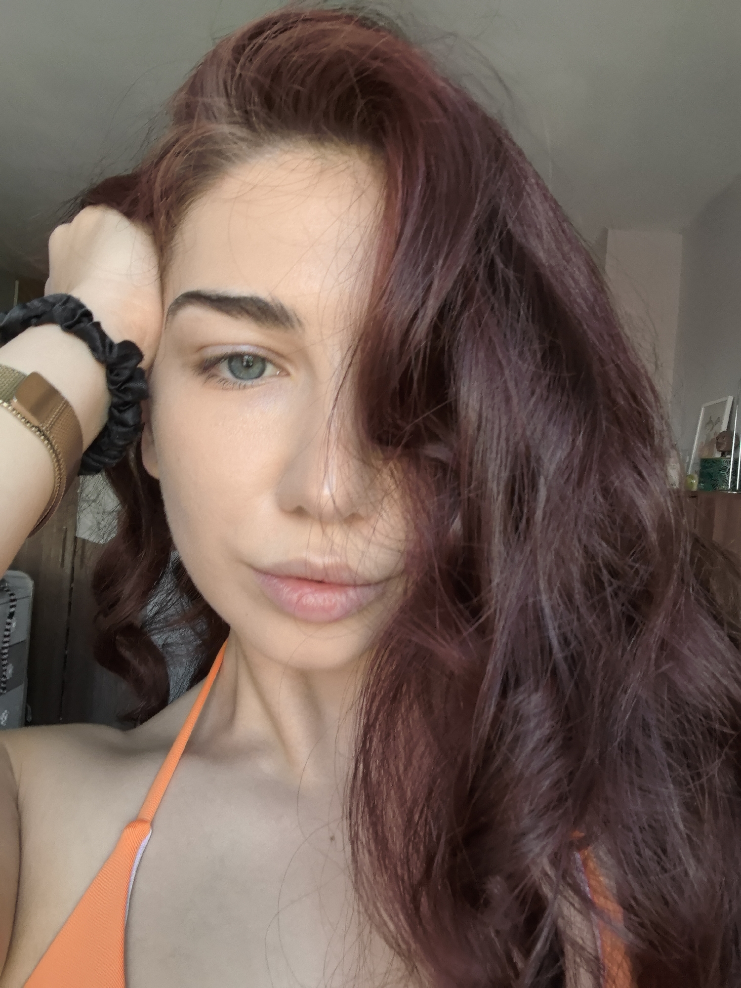 A woman with reddish-brown hair is taking a selfie indoors. She is wearing an orange bikini top and a gold bracelet on her left wrist. Her hand is resting on her head, and her hair is partially covering her face. The background appears to be a room with a wall and some furniture. There is no indication of the location being Stara Zagora, Bulgaria.