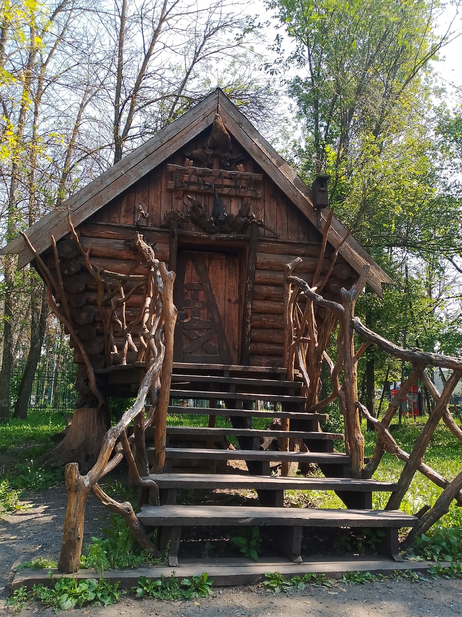 A wooden house resembling a fairy-tale dwelling sits nestled among trees in Penza, Russia. The house, constructed from rough-hewn logs, features a steeply pitched roof and a prominent wooden door. A set of wide wooden stairs with a uniquely crafted railing made of intertwined branches leads up to the entrance. The house is surrounded by lush green grass and trees, with sunlight filtering through the leaves. The overall impression is one of rustic charm and a connection to nature.