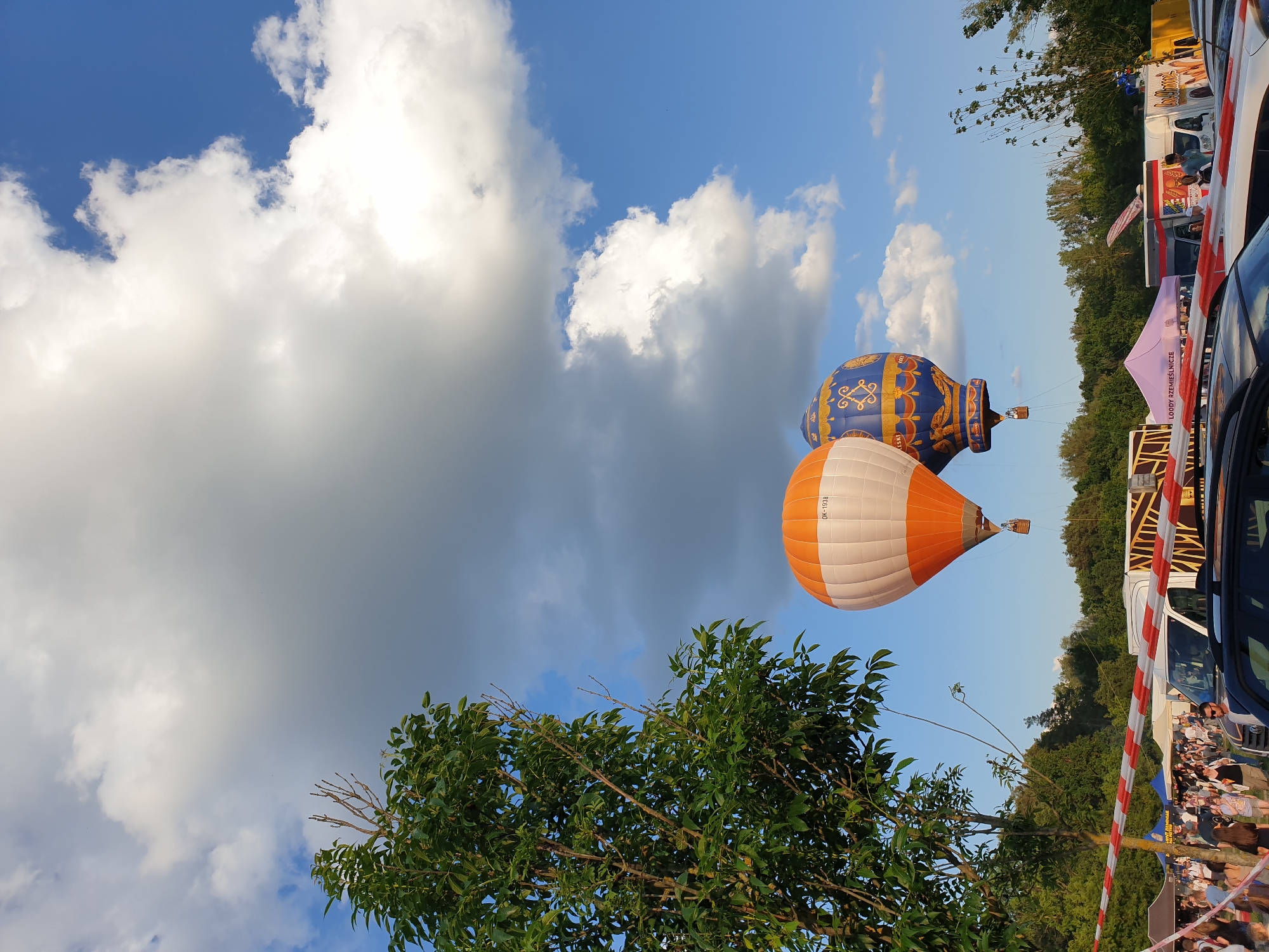 An outdoor event space under a partially cloudy blue sky is depicted, with two hot air balloons prominent in the middle ground. The left balloon is striped orange and white, displaying "OK-1938" on its side, with a visible basket beneath it. The right balloon is predominantly blue with intricate gold patterns and a basket. Both balloons are airborne, positioned above a distant treeline.
In the foreground and mid-ground, a vibrant green tree occupies the left side, its foliage extending upwards and to the right. Below the balloons, a red and white striped barrier extends horizontally, separating a crowd of people from a rear area. Numerous individuals are visible behind this barrier, observing the scene. Beyond the crowd and barrier, a collection of white vans, food trucks, and event tents are arranged. One purple tent clearly displays "LOODY RZEMIEŚLNICZE" in white text. Partial views of parked vehicles, including a dark-colored car, are visible in the immediate lower foreground, positioned behind the red and white barrier. The sky above features large, bright white cumulus clouds against a clear blue background, suggesting daylight conditions, possibly late afternoon or early evening. The overall setting is an open field or park, likely hosting a public festival or ballooning event in Unknown, Unknown.