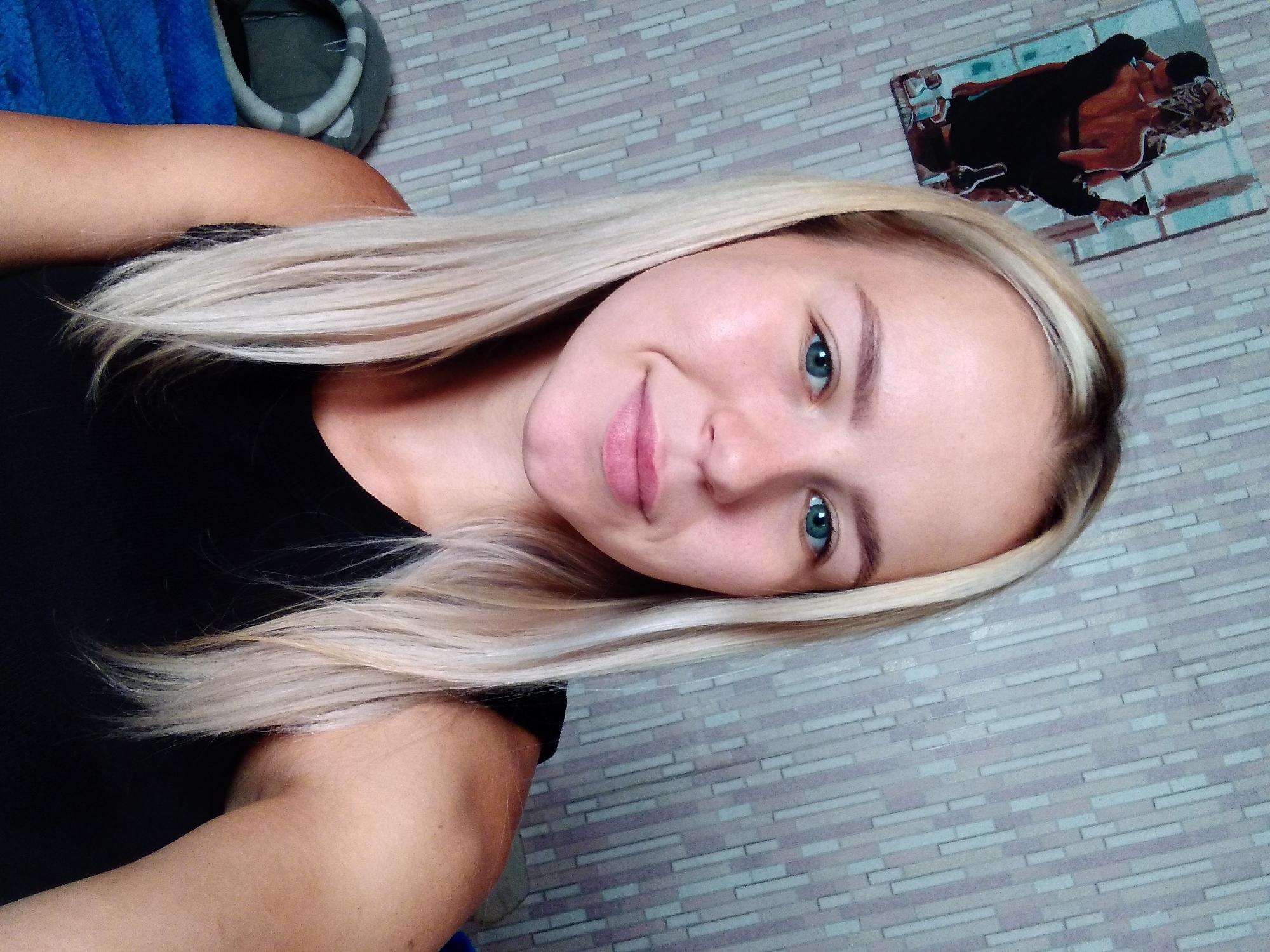 A young Caucasian woman with blonde hair and blue eyes is pictured in a selfie. She is wearing a black tank top. The background consists of a patterned wallpaper and a framed picture on the wall depicting two people. To the left of the woman, part of a pet bed and a blue blanket are visible. The woman is likely indoors, in a residential setting in Radisne, Ukraine.