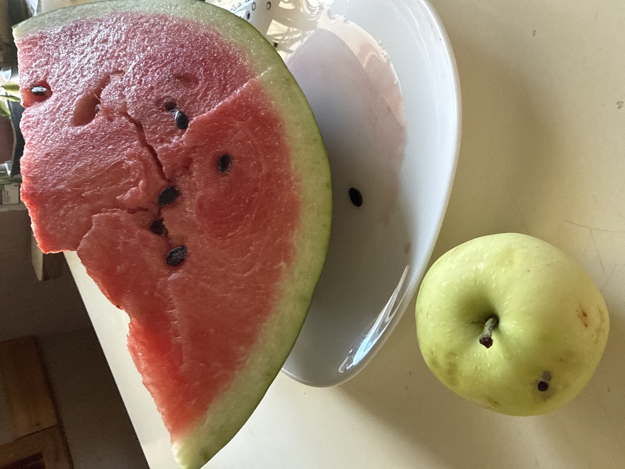 A slice of watermelon sits on a white plate with a few seeds and a pink stain. To the left of the plate is a green apple with a brown stem and several dark spots. The scene appears to be indoors on a light-colored surface. There is no indication of the location being in Okhtyrka, Ukraine.