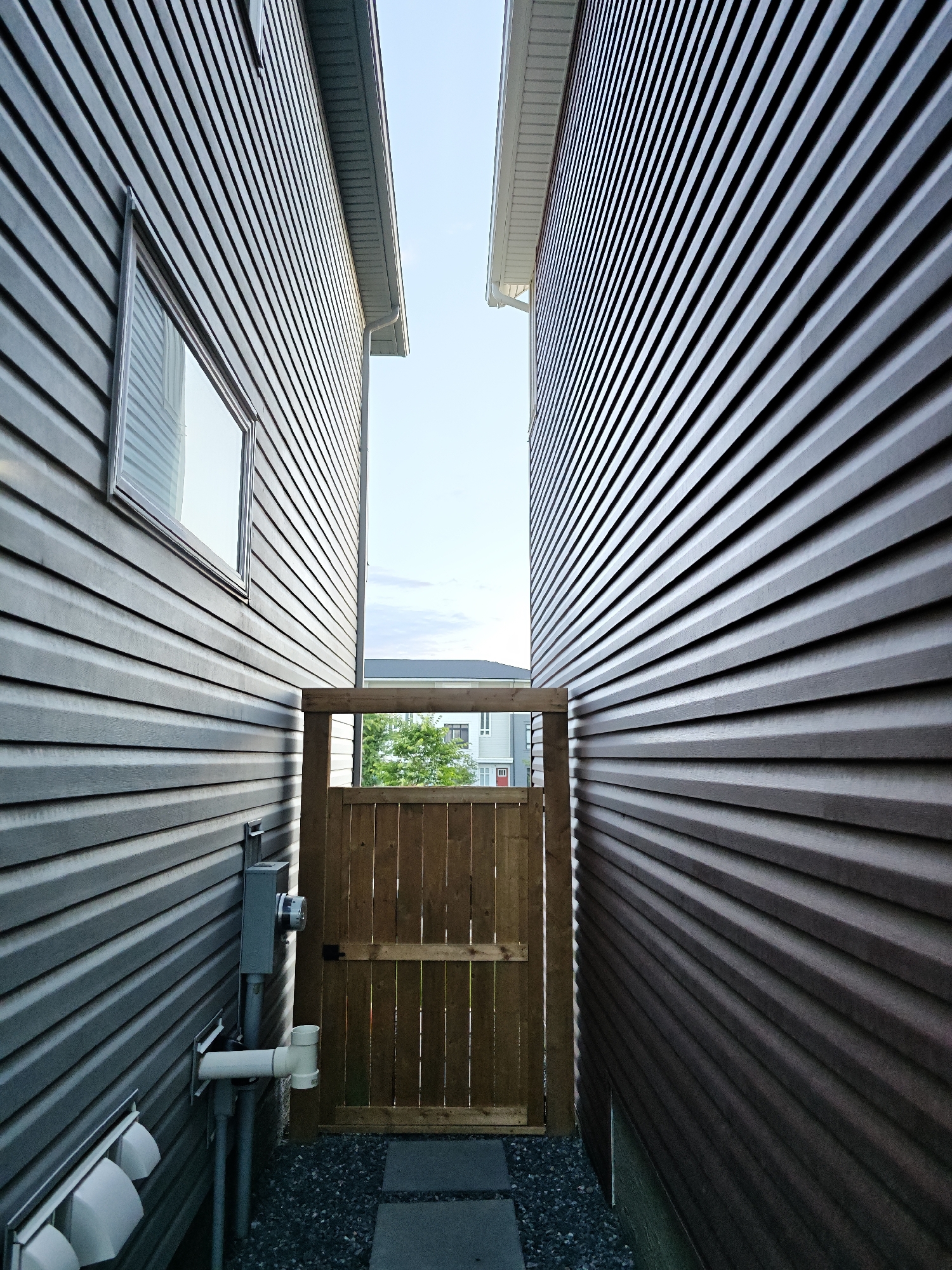 This is an exterior, low-angle, wide shot of a narrow passage between two residential buildings with gray horizontal siding. The passage leads to a wooden gate. The sky is visible between the buildings at the top of the frame. On the left building, there is a window and some utility boxes and pipes. The ground in the passage is covered with dark gravel and has two large gray pavers. In the background, through the gate, there are trees and parts of other houses visible. The scene appears to be in a suburban area.