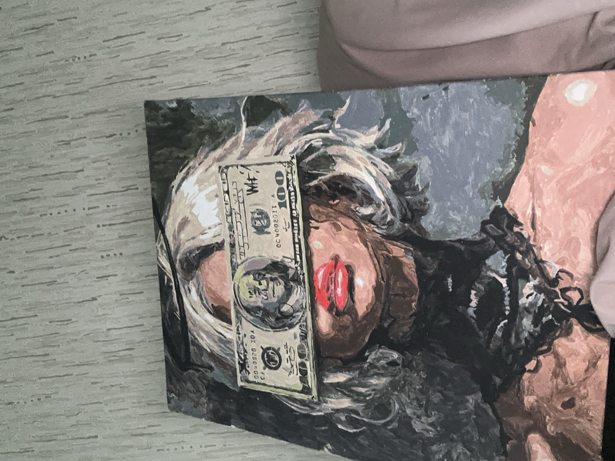 A painting on canvas depicts a person's face, partially obscured by a one-hundred dollar bill. The person has blonde hair, visible red lips, and is partially covered with black cloth or material. The painting's background is a mix of grey and black hues. The image was taken indoors on what appears to be a light-colored, textured surface, such as a table or countertop. The location is Высокий, Russia.