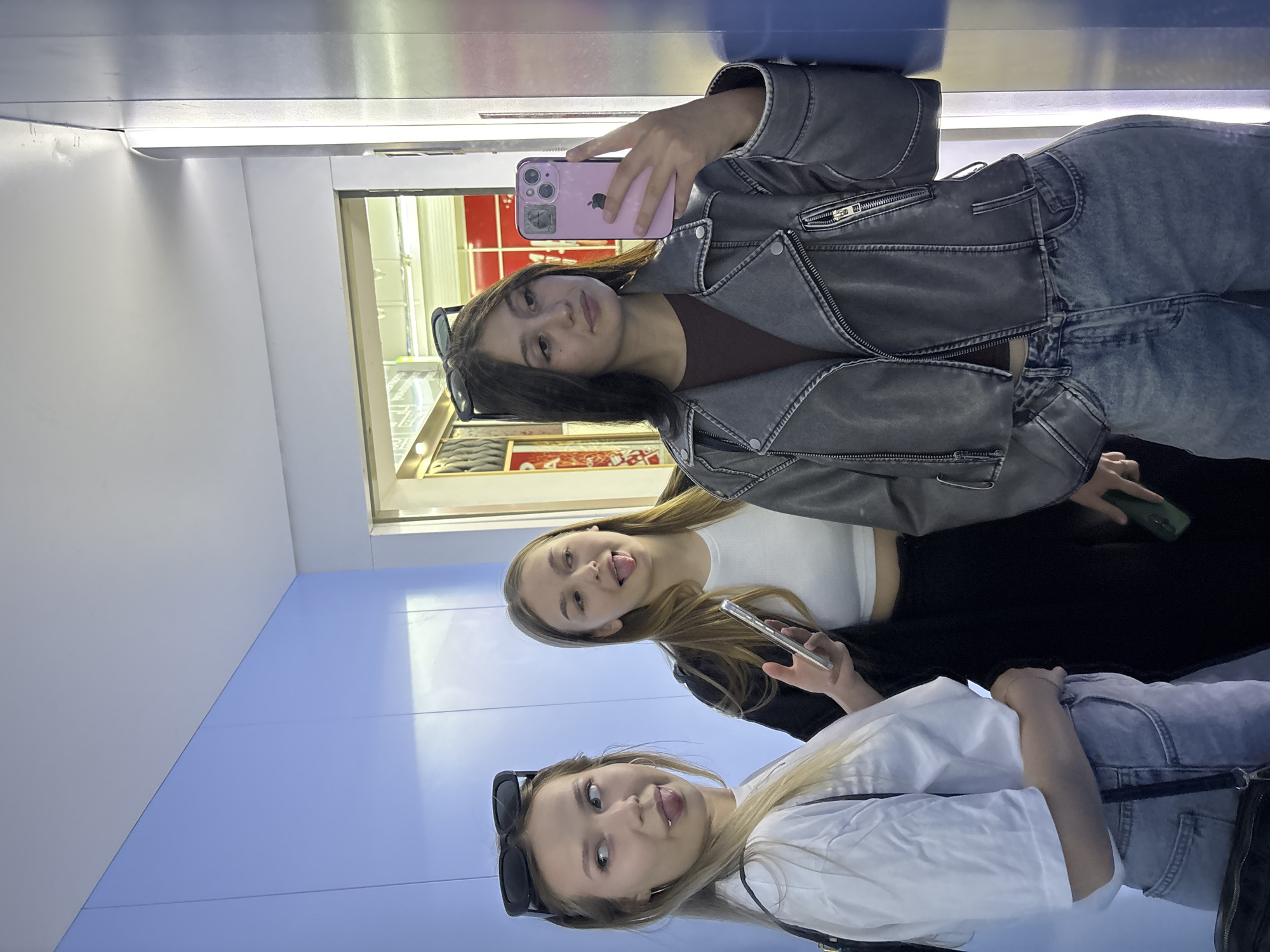 Three young women are posing for a photo in what appears to be a brightly lit interior, possibly an elevator or a narrow hallway. The walls are a vibrant light blue, and a vertical strip of metallic material runs along the right side of the frame.
The woman on the left, with blonde hair and sunglasses perched on her head, is making a face with her tongue sticking out. She is wearing a white t-shirt and light-wash jeans. A black strap from a bag crosses her chest.
The woman in the middle, with long blonde hair, is also sticking her tongue out and holding a smartphone. She has on a white cropped top and a black jacket over dark pants.
The woman on the right, with dark hair, is holding a pink smartphone and taking a selfie. She is wearing a grey moto jacket over a dark top and light-wash jeans. She has sunglasses on her head.
The background shows a glimpse of a retail environment with displays and lighting. There is no direct indication of the city of Chaikovsky, Russia, within the image itself; this information is provided externally.