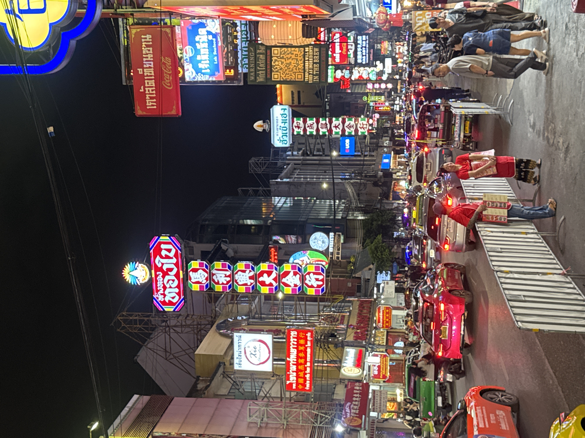 This is a photograph taken at night on a bustling street in Bangkok, Thailand, specifically in the Chinatown area. The image captures the vibrant atmosphere with a high density of colorful neon signs in both Thai and Chinese characters. Several cars and motorcycles are driving down the street, indicating traffic. People are walking on the sidewalk, some wearing casual attire. A couple of vendors stand near the edge of the sidewalk, possibly selling items or menus. Various storefronts and restaurants are visible behind the signs.